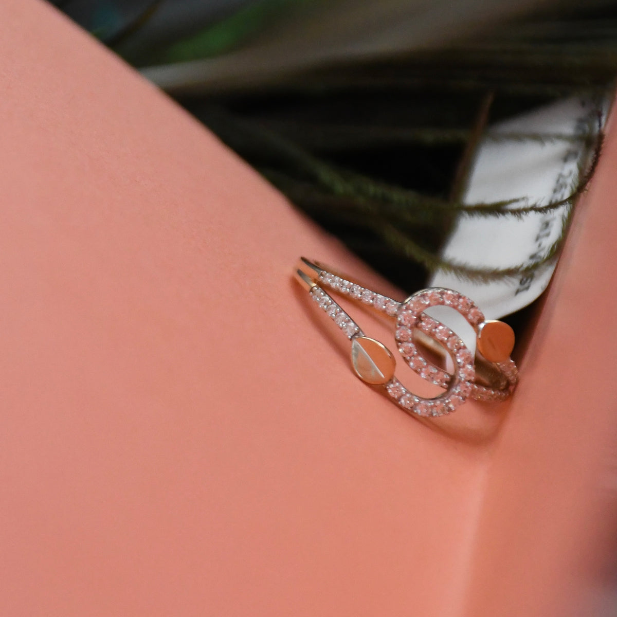 A close-up of a rose gold ring with a double knot design, embellished with small clear pavé stones and a central oval inlay, resting on a peach-colored surface.