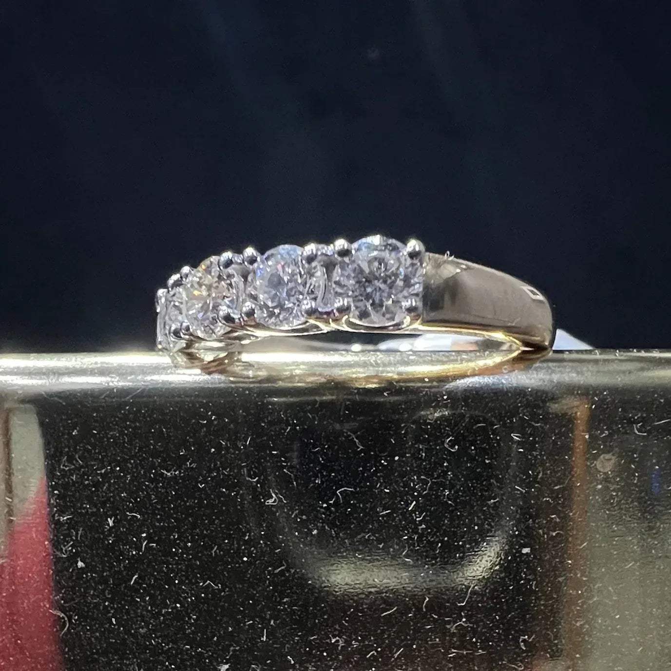 A close-up image of a white gold half eternity ring featuring a row of round brilliant-cut diamonds set in prongs, displayed on a reflective surface against a dark background, highlighting the sparkle and detailed craftsmanship of the ring.
