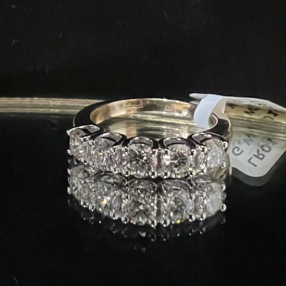 A close-up image of a white gold half eternity ring featuring a row of round brilliant-cut diamonds set in prongs, displayed on a reflective surface against a dark background, highlighting the sparkle and detailed craftsmanship of the ring.