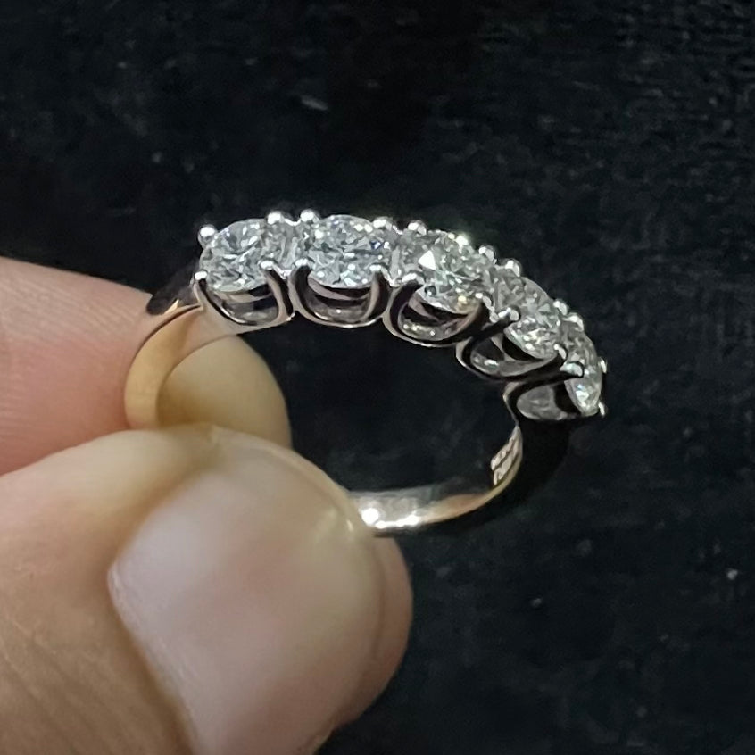 A close-up of a white gold half eternity ring featuring a row of round brilliant-cut diamonds set in prongs, held between fingers against a dark background, highlighting the sparkle and detailed craftsmanship of the ring.