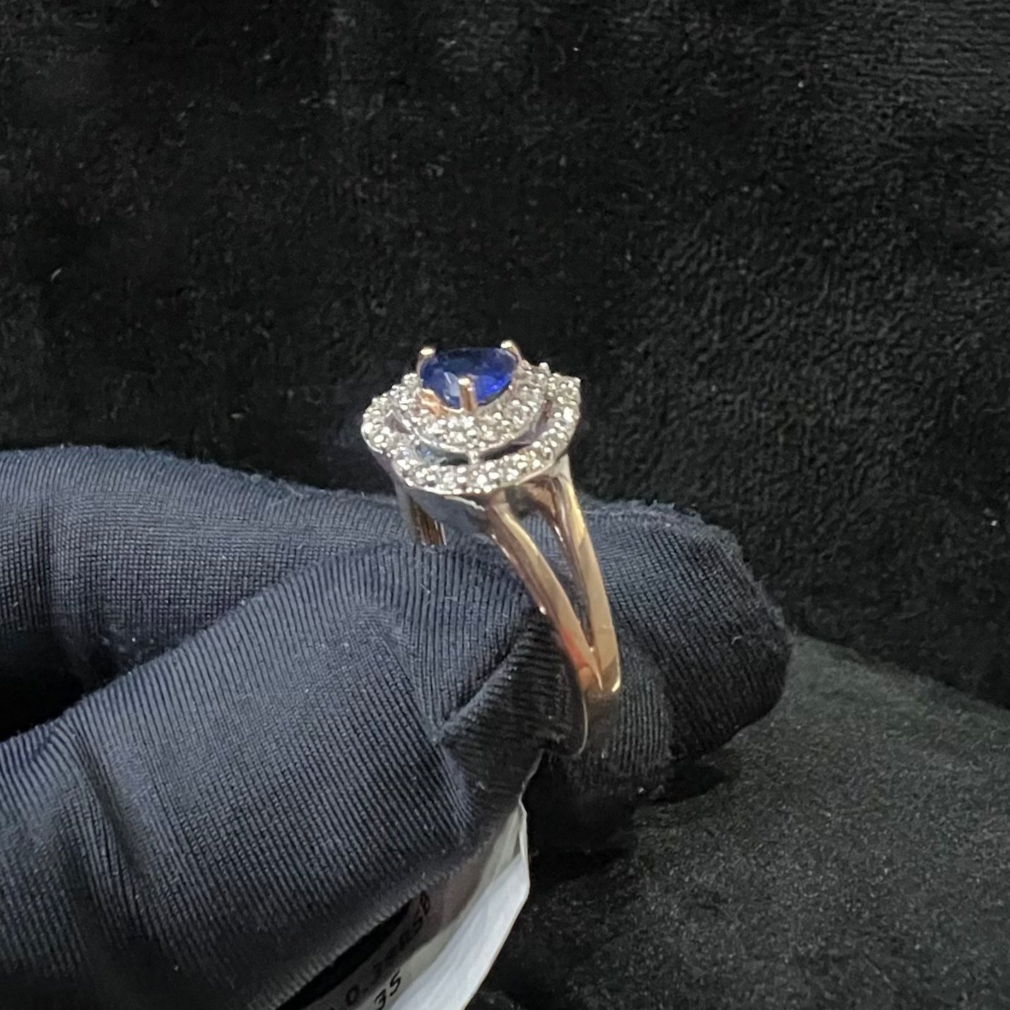 A gold ring with a central blue semi-precious stone, surrounded by smaller white diamonds, held in a gloved hand.