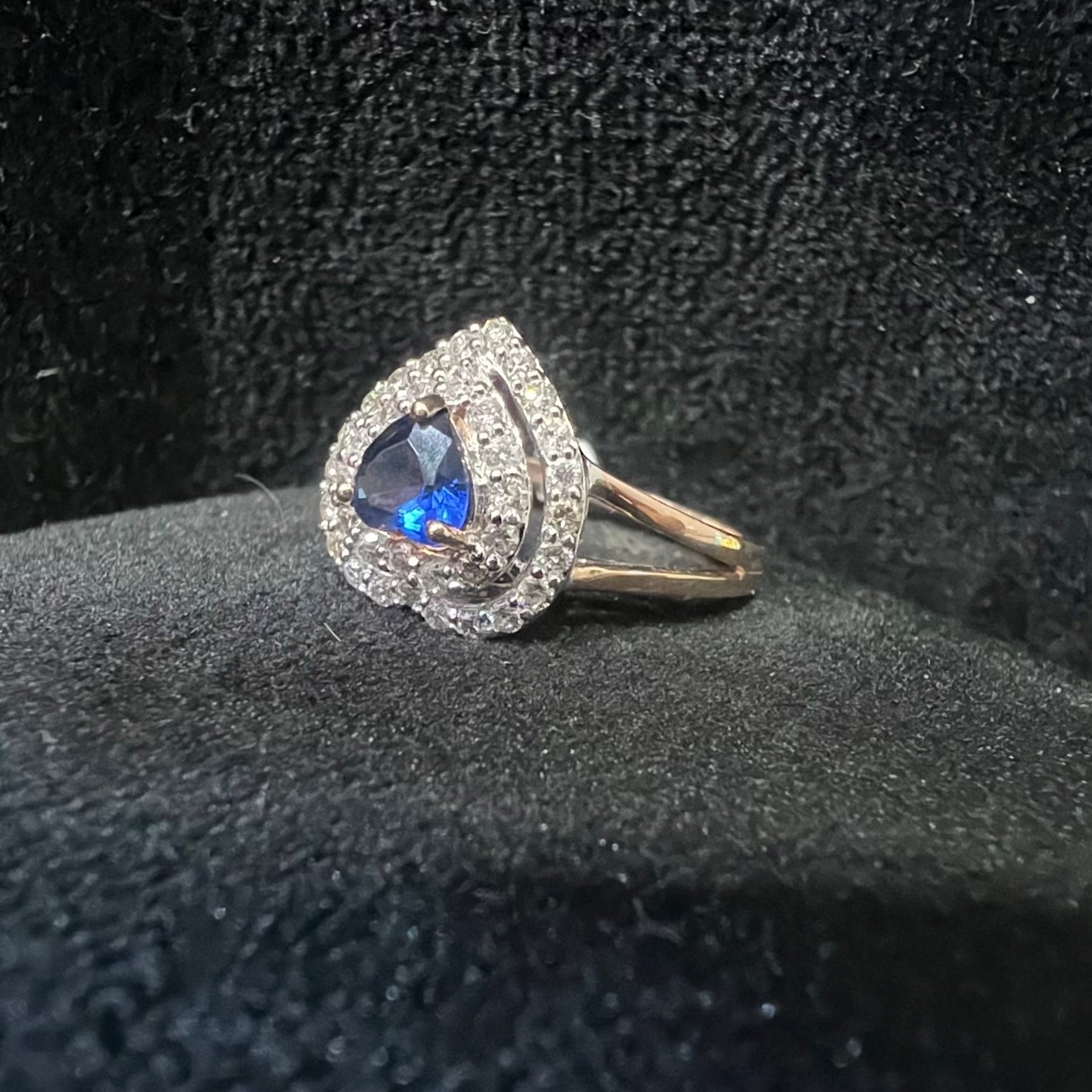 A gold ring with a central blue semi-precious stone, surrounded by smaller white diamonds, displayed on black rough surface.