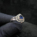 A gold ring with a central blue semi-precious stone, surrounded by smaller white diamonds, held in a gloved hand.