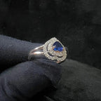 A gold ring with a central blue semi-precious stone, surrounded by smaller white diamonds, held in a gloved hand.