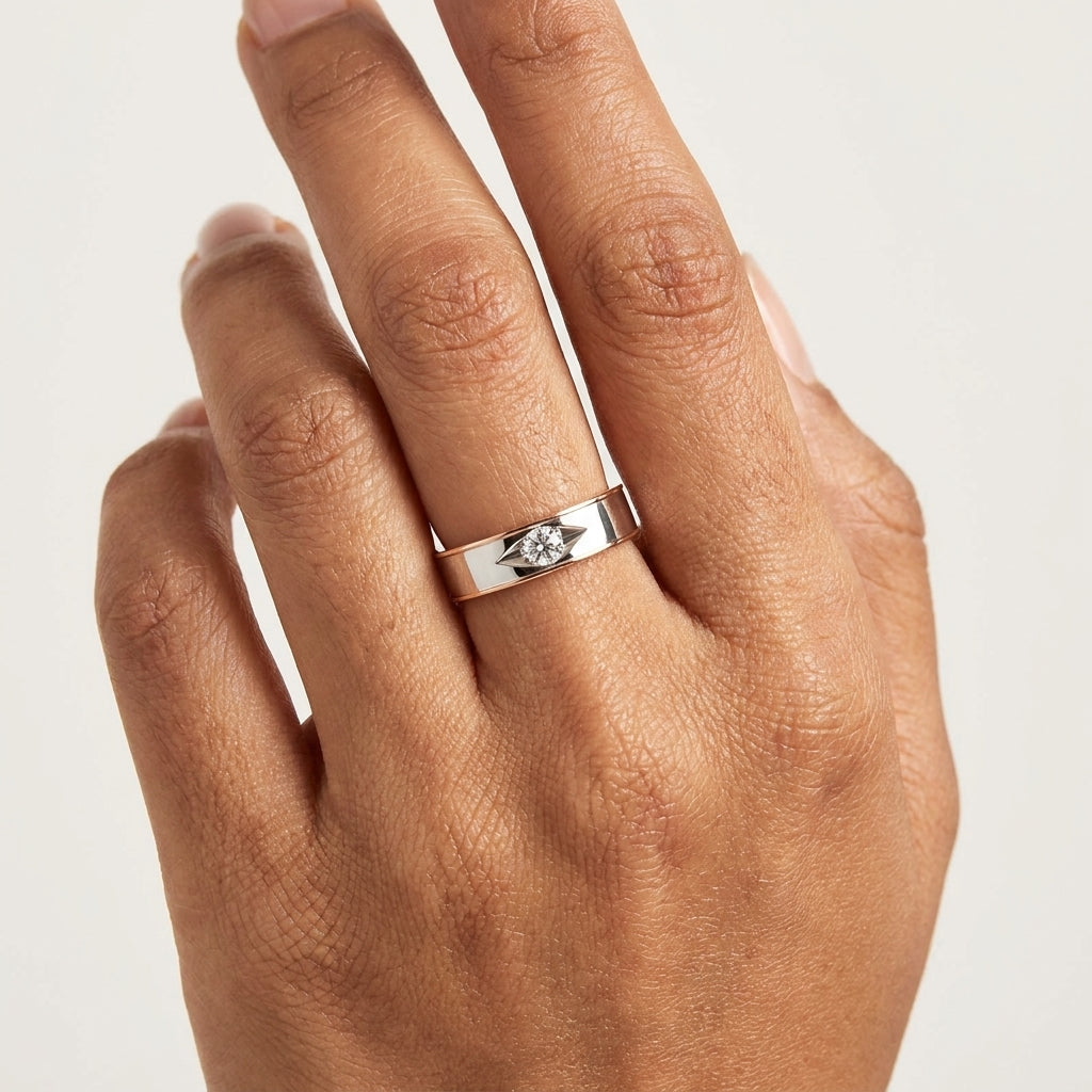 A close-up of solitaire diamond engagement ring with a gold band featuring a single diamond at the top center, set in a white and rose gold two-tone design worn in a hand's finger against a white background.