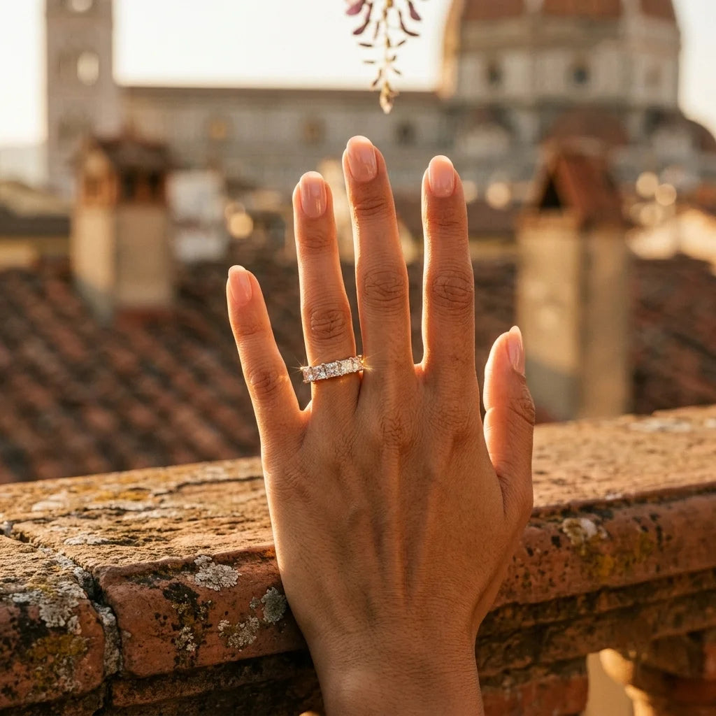 A woman's hand resting on a stone terrace, wearing a yellow gold natural diamond multi-row ring, with a softly blurred historic cityscape illuminated by sunset.
