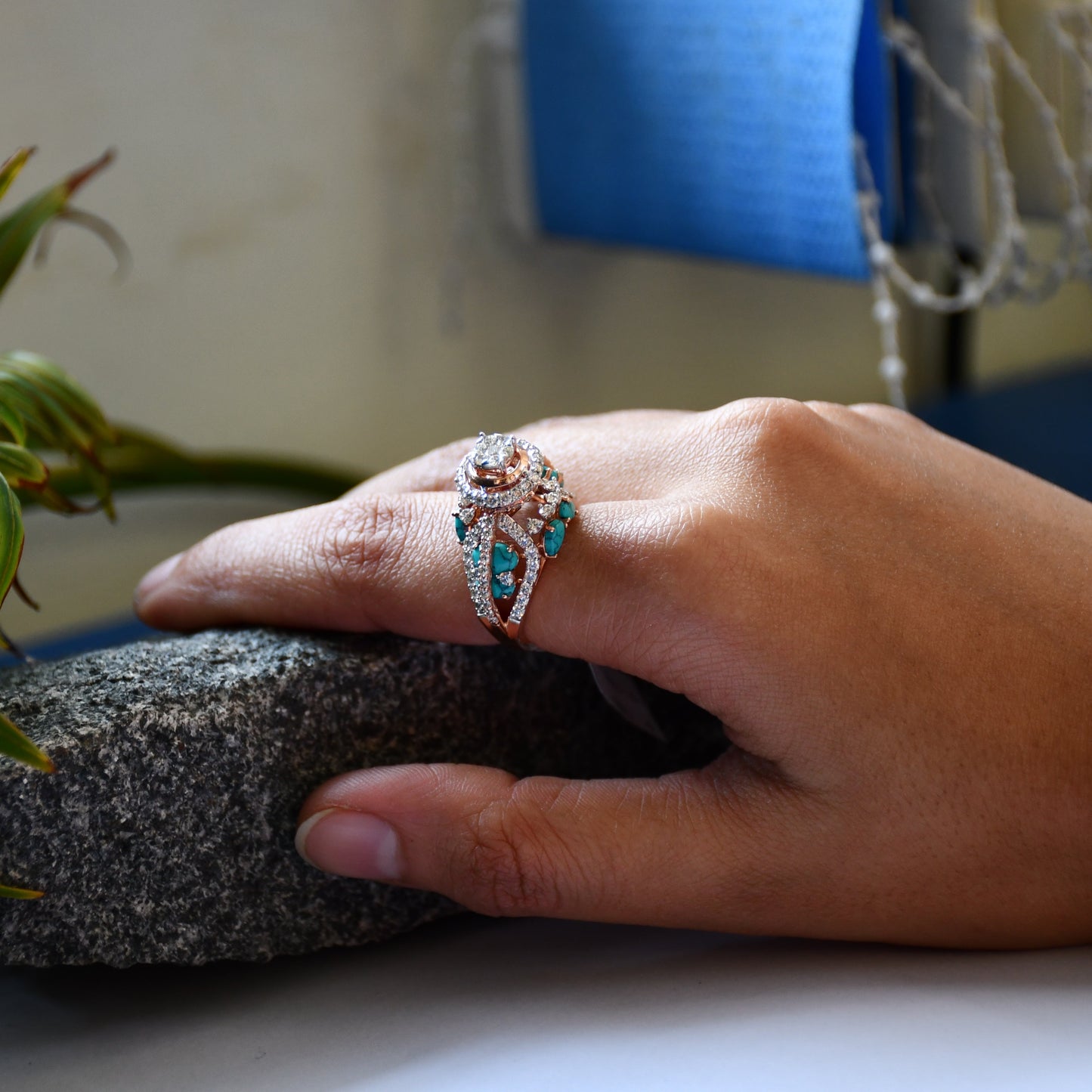A woman's hand on a rock stone displaying a rose gold diamond ring with a central brilliant cut diamonds and smaller color stones around it.