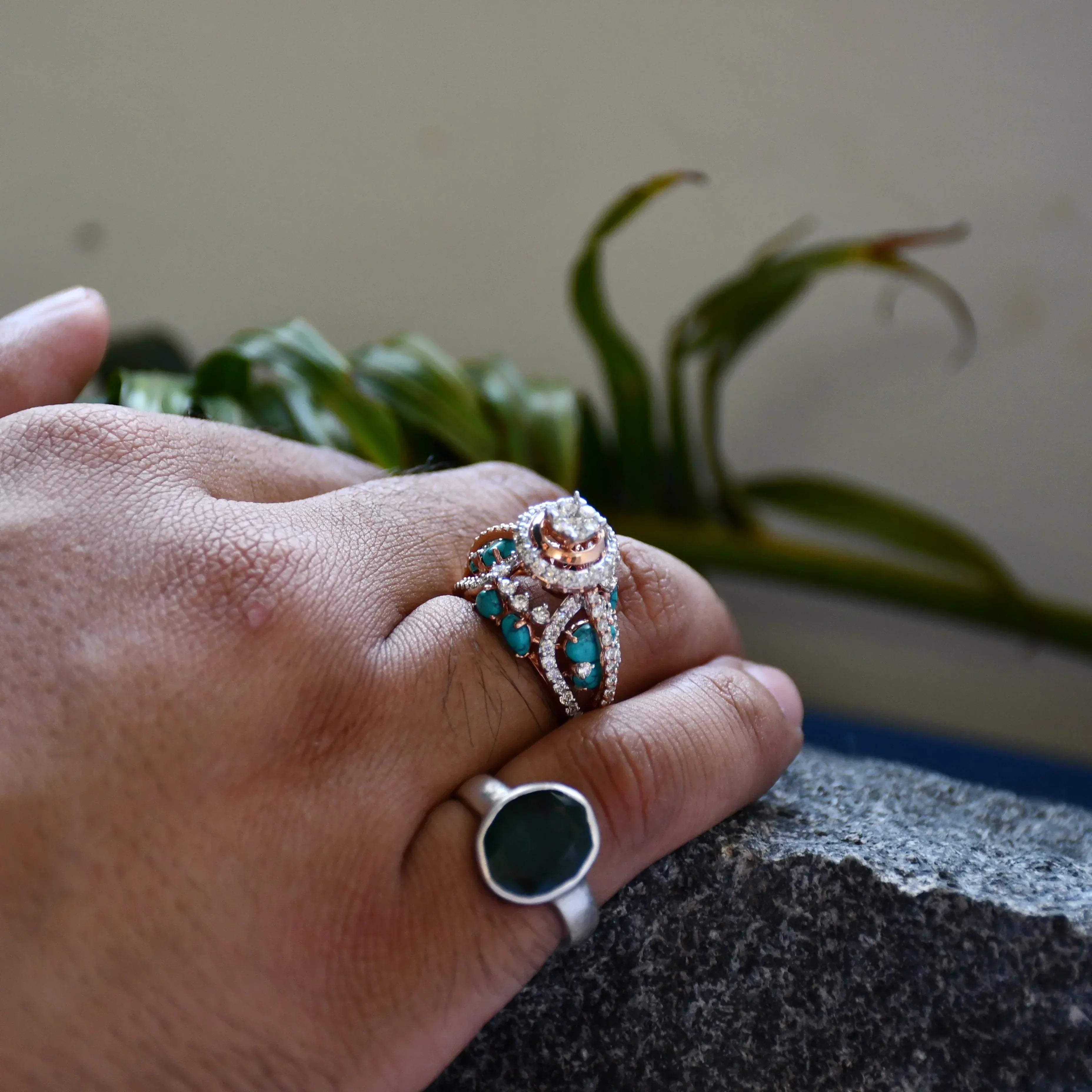 A woman's hand on a rock stone displaying a rose gold diamond ring with a central brilliant cut diamonds and smaller color stones around it.