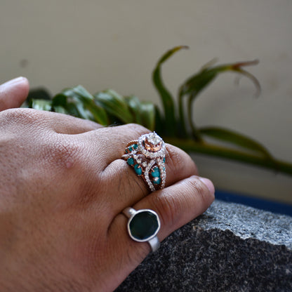 A woman's hand on a rock stone displaying a rose gold diamond ring with a central brilliant cut diamonds and smaller color stones around it.