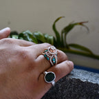 A woman's hand on a rock stone displaying a rose gold diamond ring with a central brilliant cut diamonds and smaller color stones around it.