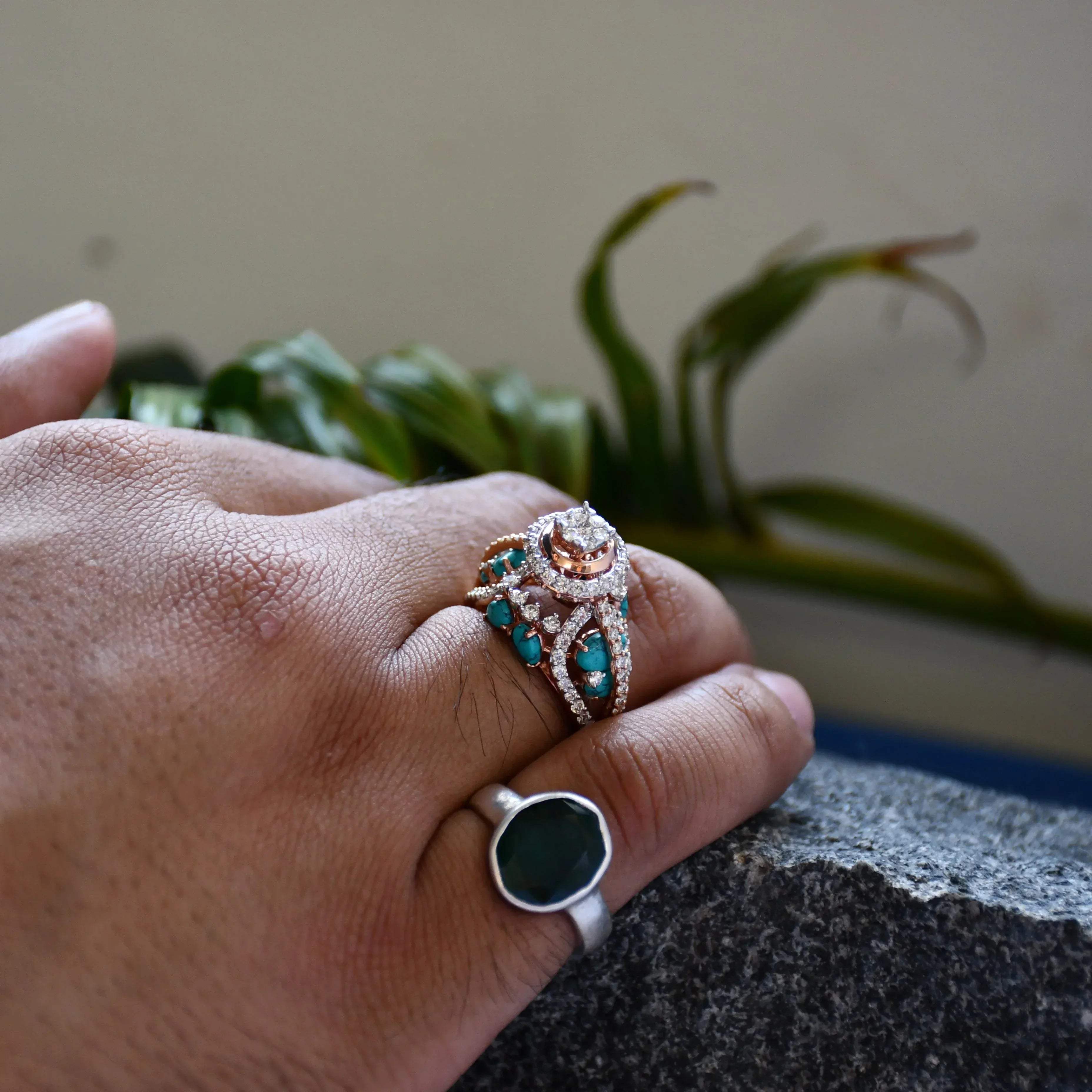 A woman's hand on a rock stone displaying a rose gold diamond ring with a central brilliant cut diamonds and smaller color stones around it.
