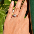 A person's hand holding a 14kt gold band ring with a round brilliant cut diamond centerpiece, set on a leafy background.