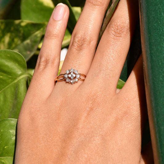 A person's hand holding a 14kt gold band ring with a round brilliant cut diamond centerpiece, set on a leafy background.