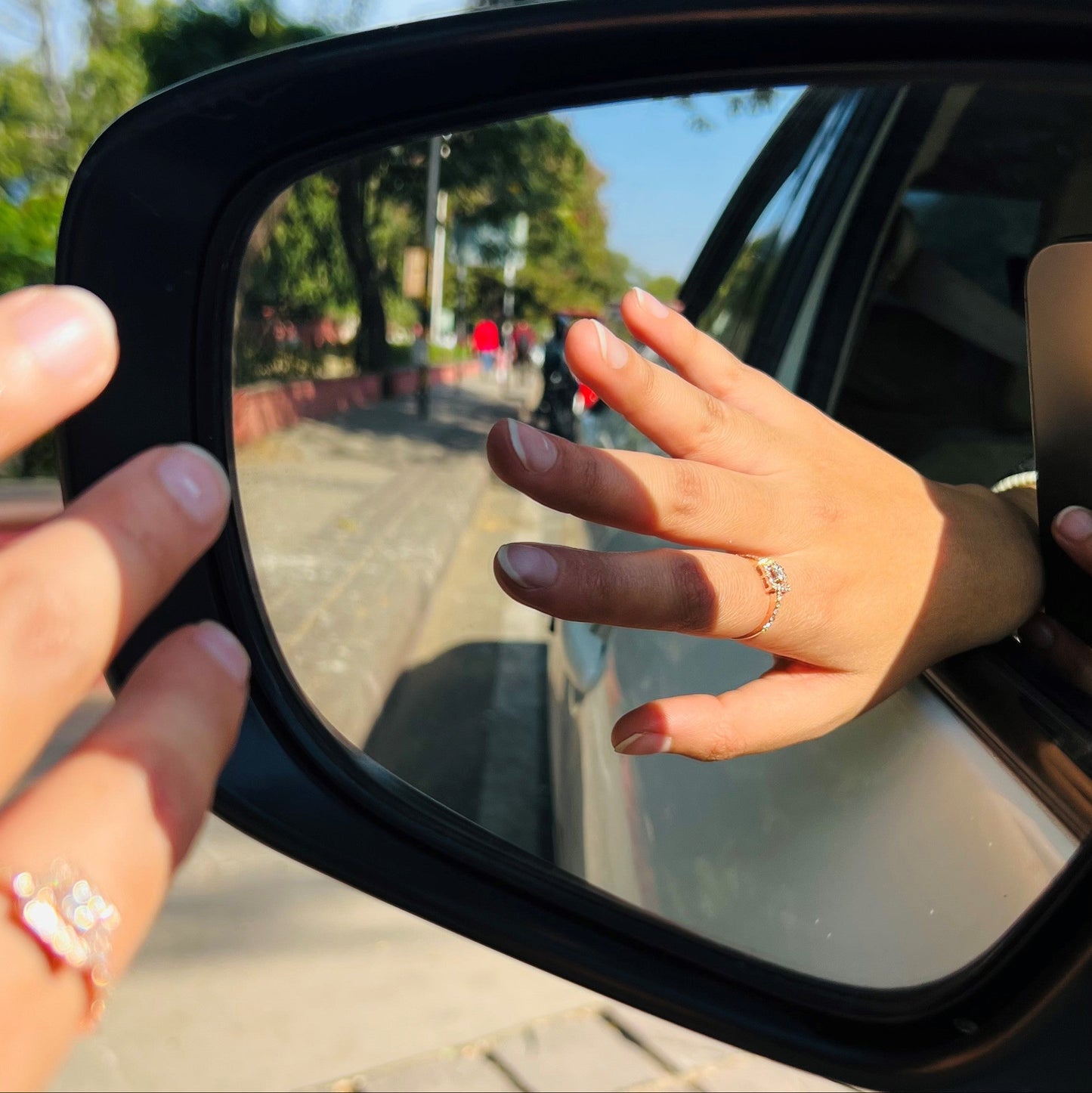 A 14K gold ring with a central square-shaped setting holding a sparkling, multicolored diamond, surrounded by smaller round diamonds set worn by a hand that is showing ring in car's mirror.