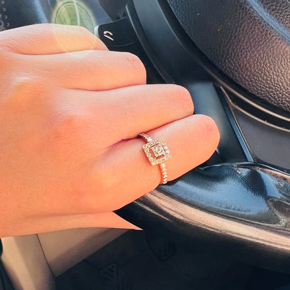 A 14K gold ring with a central square-shaped setting holding a sparkling, multicolored diamond, surrounded by smaller round diamonds set worn by a hand that is holding car's steering.