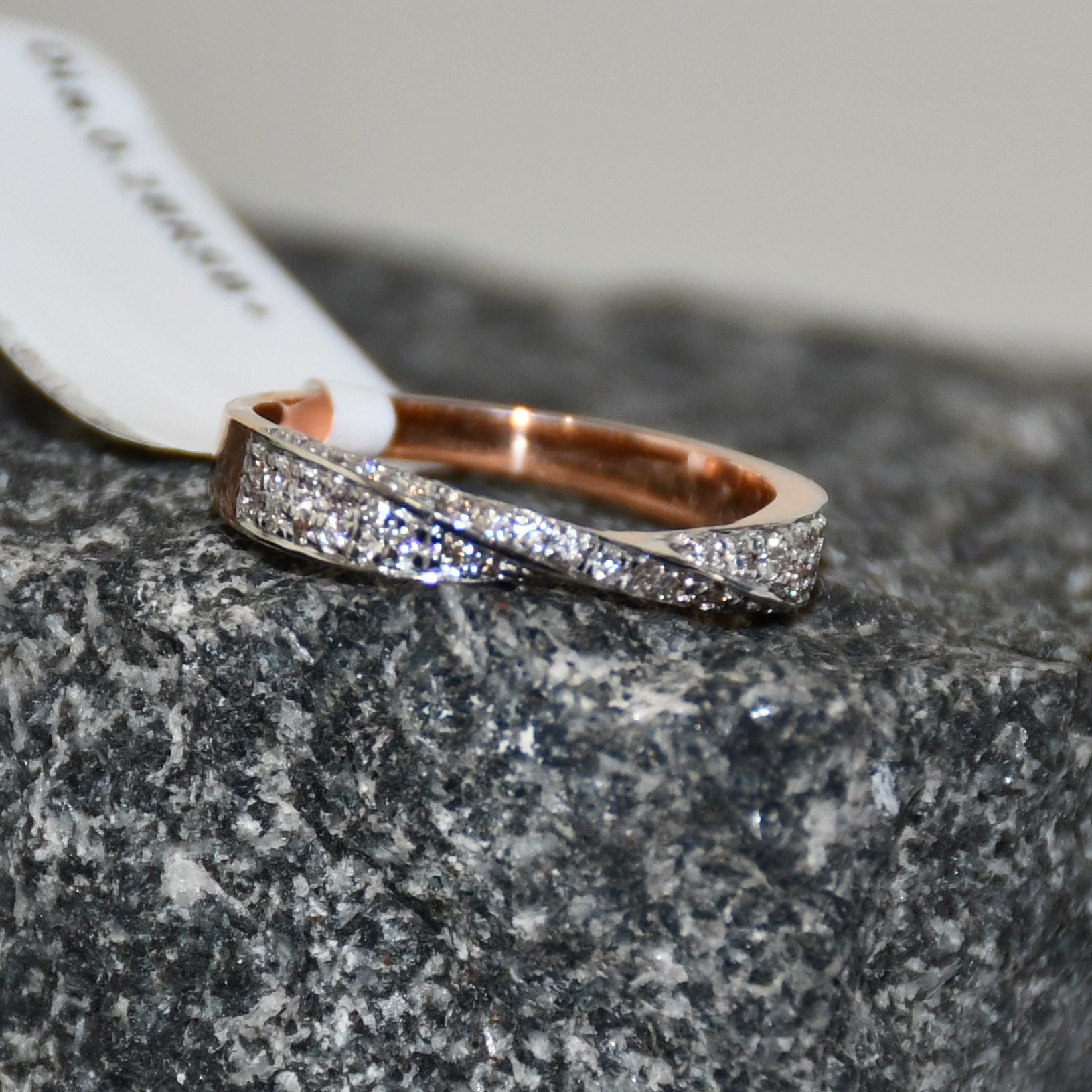 Elegant two-tone diamond ring featuring intertwined bands of rose gold and white gold, adorned with small round-cut diamonds, displayed on rough textured surface.