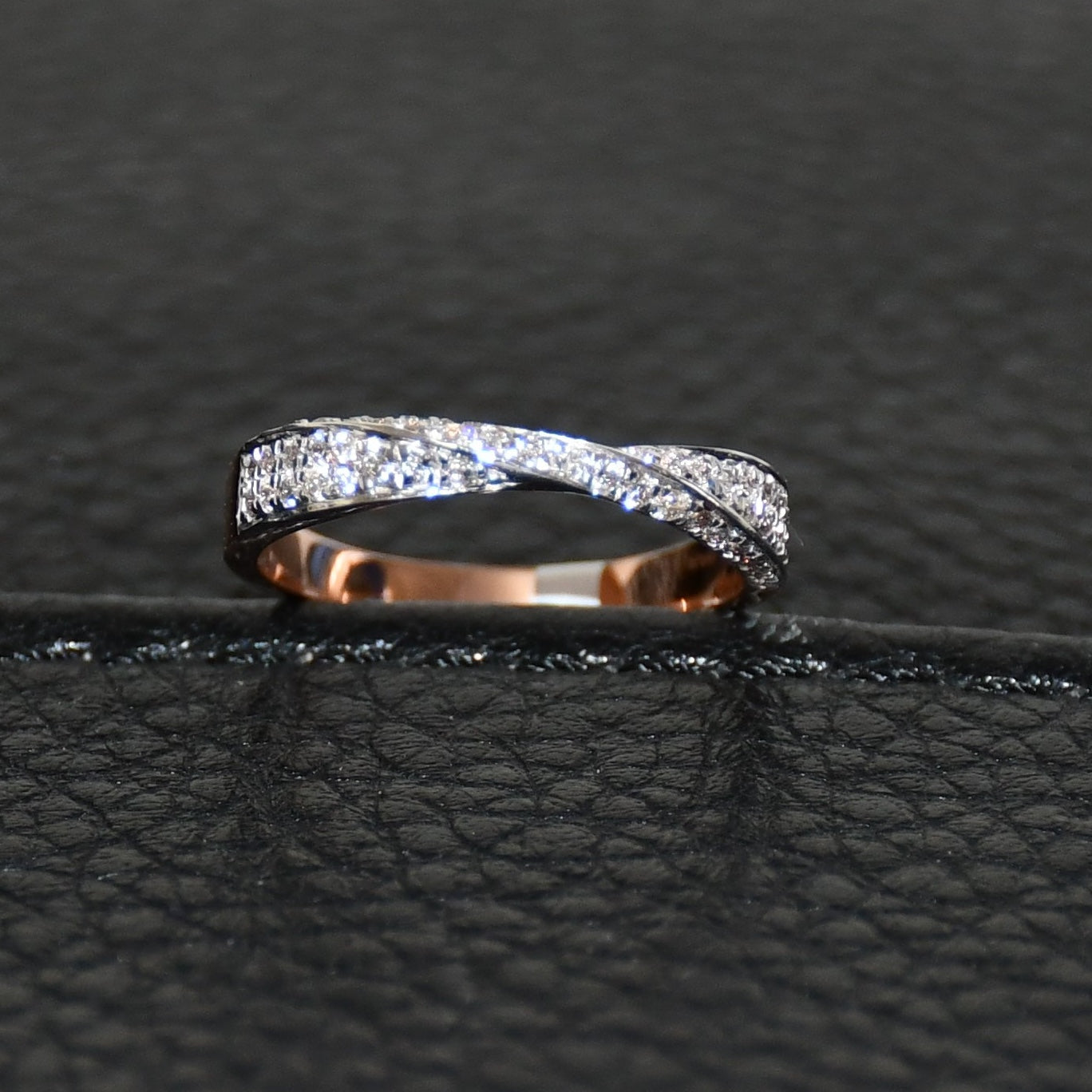 Elegant two-tone diamond ring featuring intertwined bands of rose gold and white gold, adorned with small round-cut diamonds, displayed on black leather background.