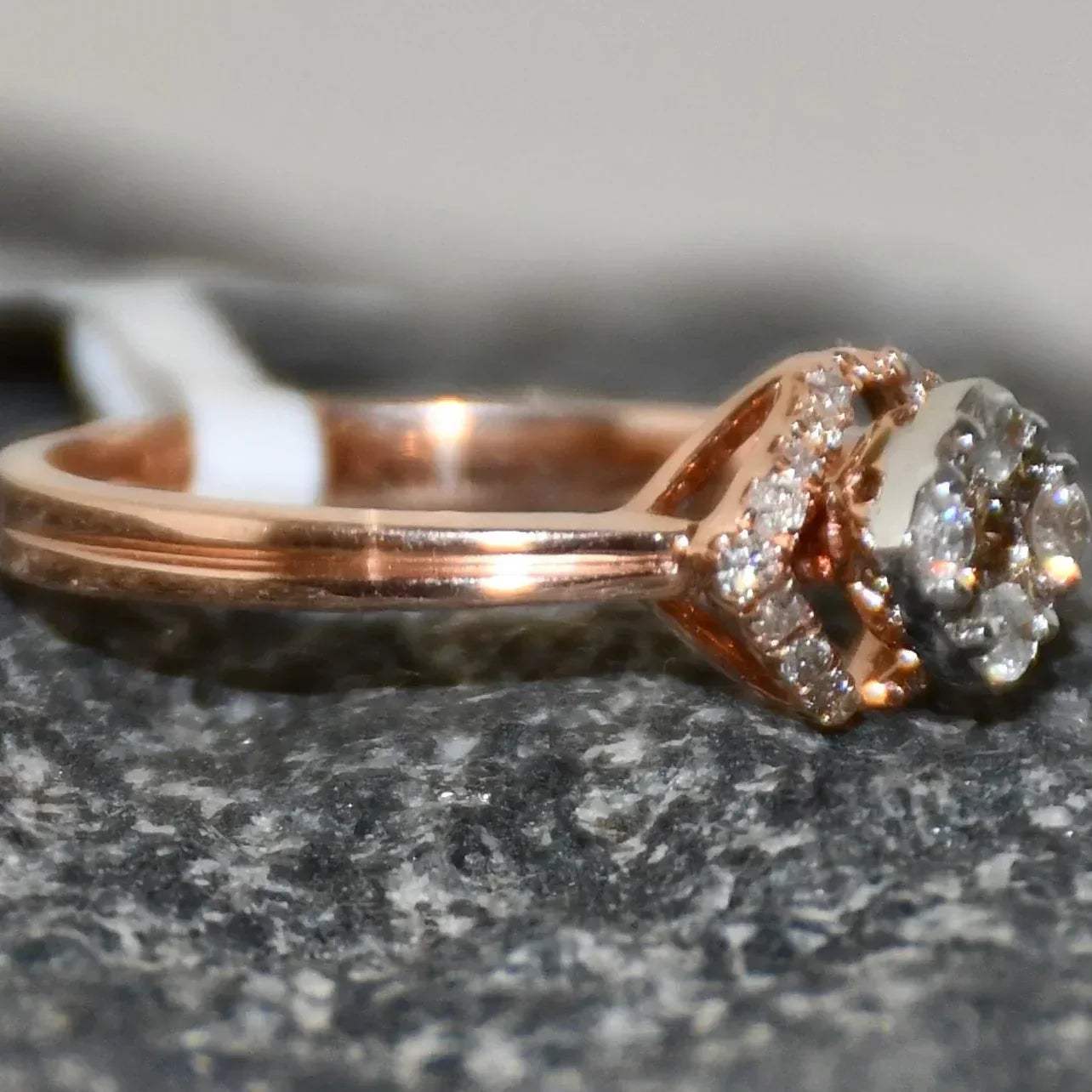 A gold ring with a central diamond surrounded by smaller diamonds set in a solid gold band is displayed on a rough & textured surface.