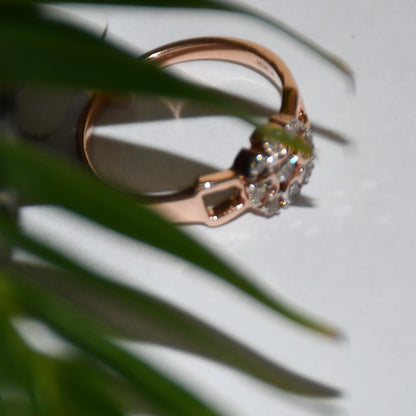 A 14K gold diamond band ring with a central row of diamonds and smaller surrounding diamonds set in a gold band displayed on a white surface and behind blur green leaves.