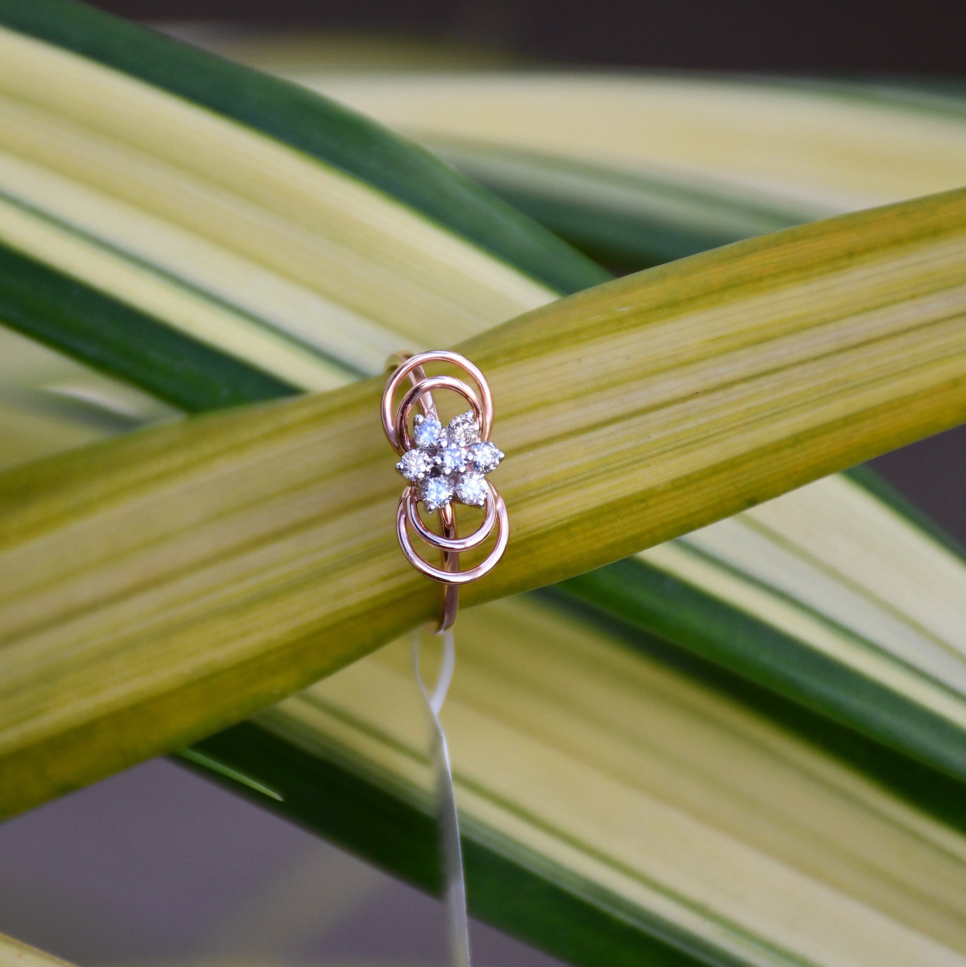 Delicate rose gold ring featuring a floral cluster of round diamonds at the center, showcased on green and yellow variegated leaves.