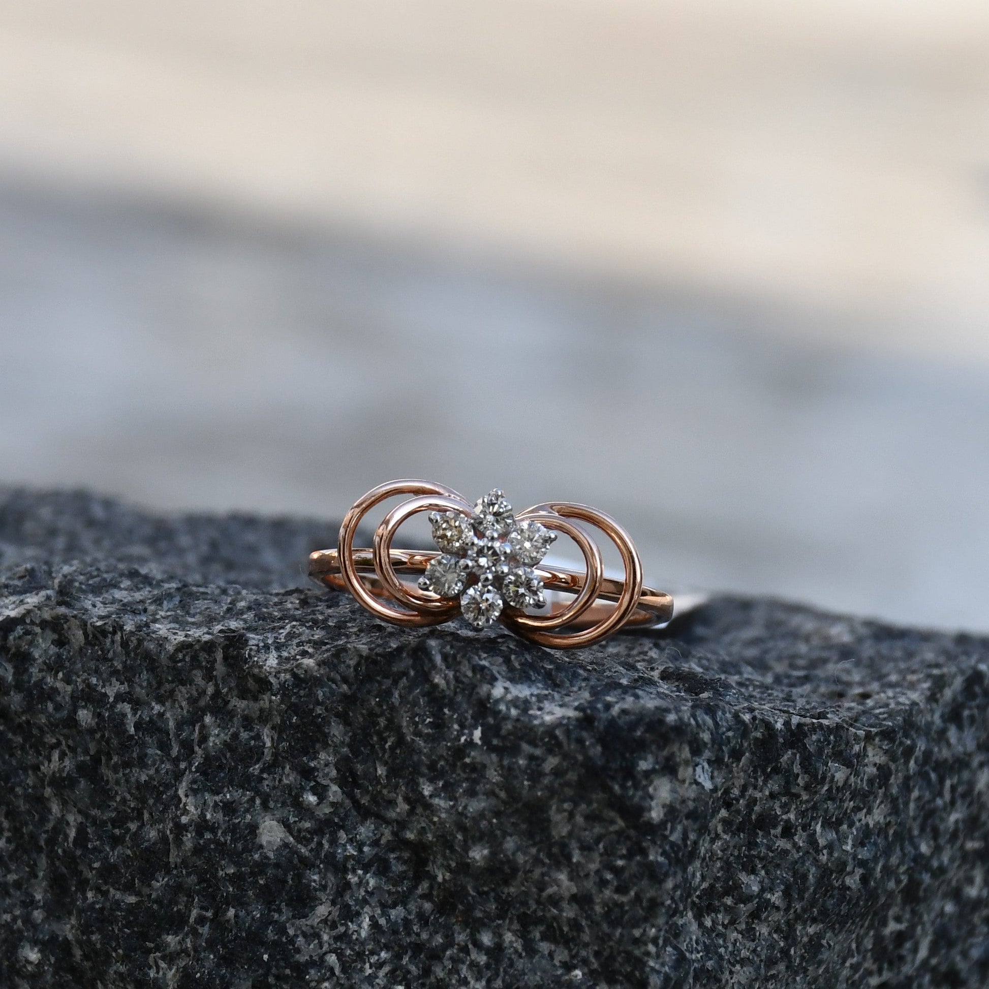 Delicate rose gold ring featuring a floral cluster of round diamonds at the center, showcased on a rough & textured surface.