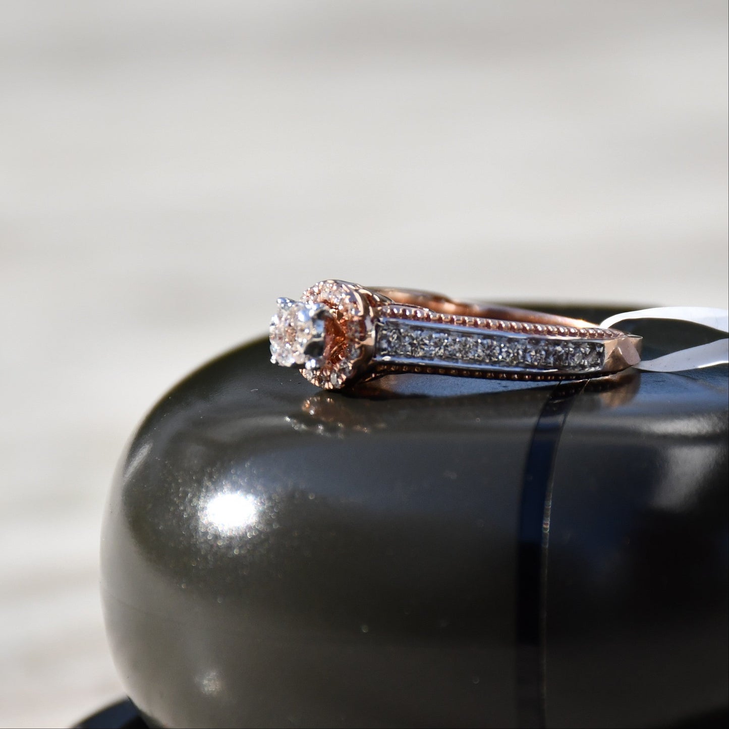 Close-up of a gold diamond ring displayed on a black smooth surface, featuring a round-cut center diamond surrounded by a delicate halo and a tapered band accented with small pavé-set diamonds and fine milgrain detailing.