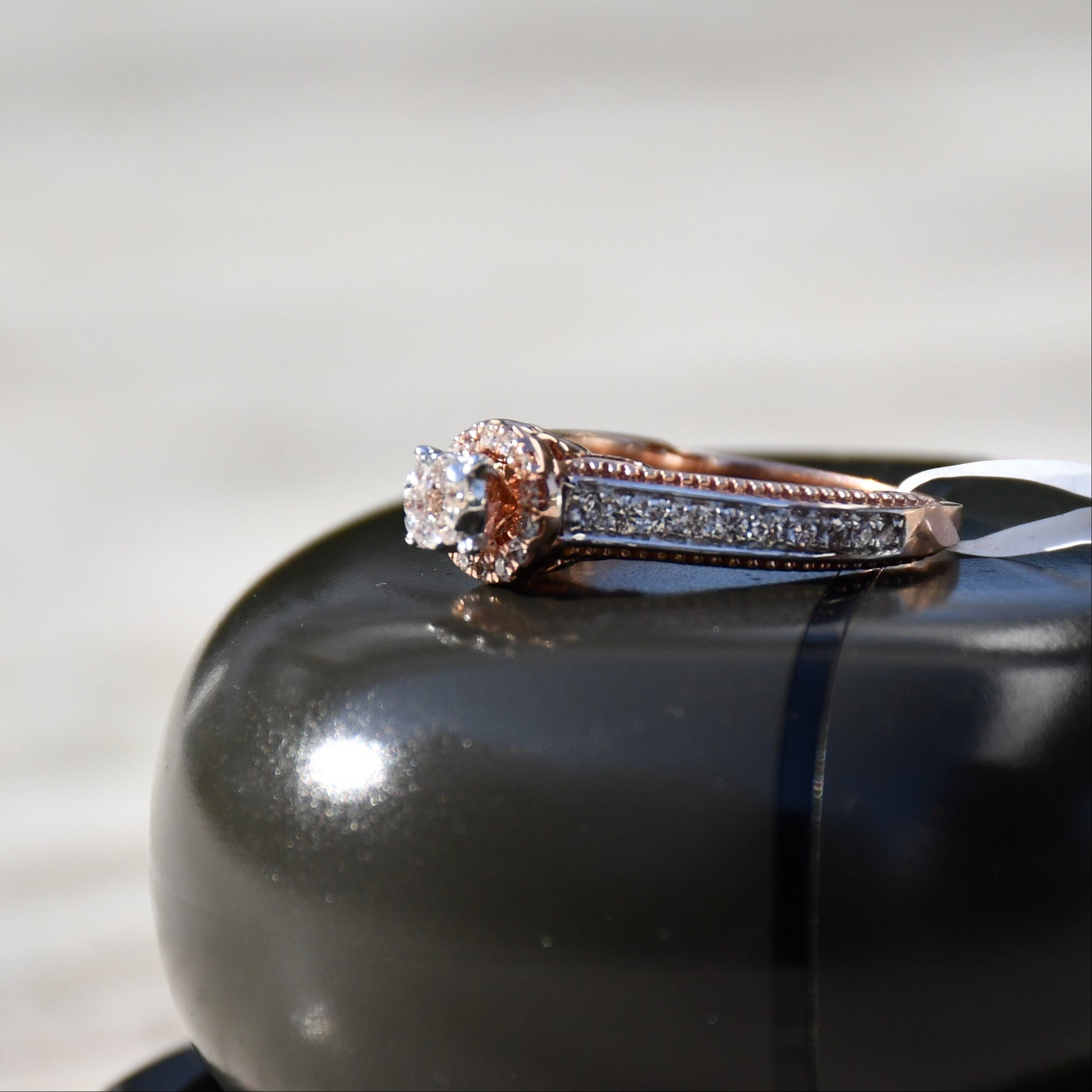Close-up of a gold diamond ring displayed on a black smooth surface, featuring a round-cut center diamond surrounded by a delicate halo and a tapered band accented with small pavé-set diamonds and fine milgrain detailing.