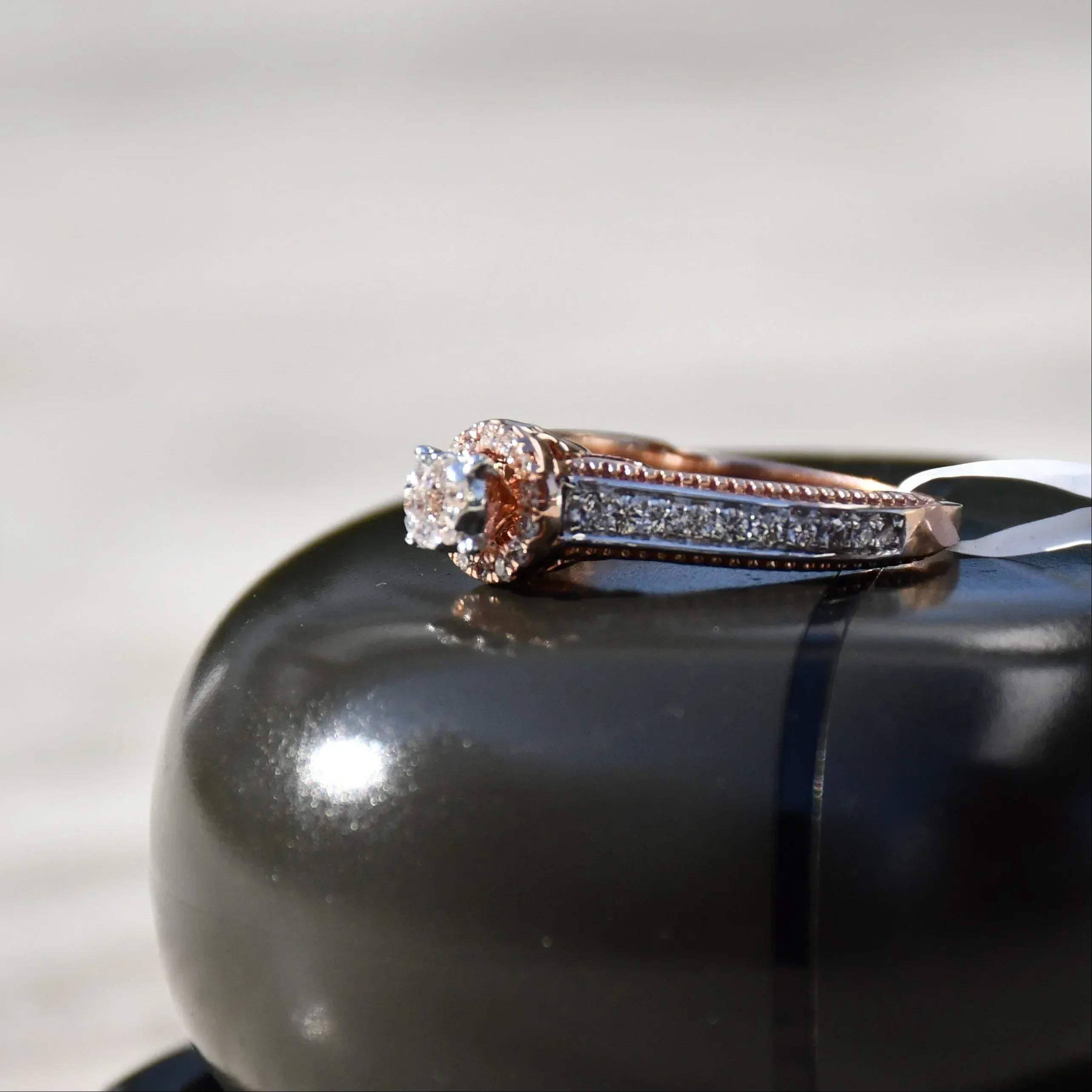 Close-up of a gold diamond ring displayed on a black smooth surface, featuring a round-cut center diamond surrounded by a delicate halo and a tapered band accented with small pavé-set diamonds and fine milgrain detailing.