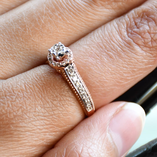 Close-up of a gold diamond ring worn on a finger, featuring a round-cut center diamond surrounded by a delicate halo and a tapered band accented with small pavé-set diamonds and fine milgrain detailing.