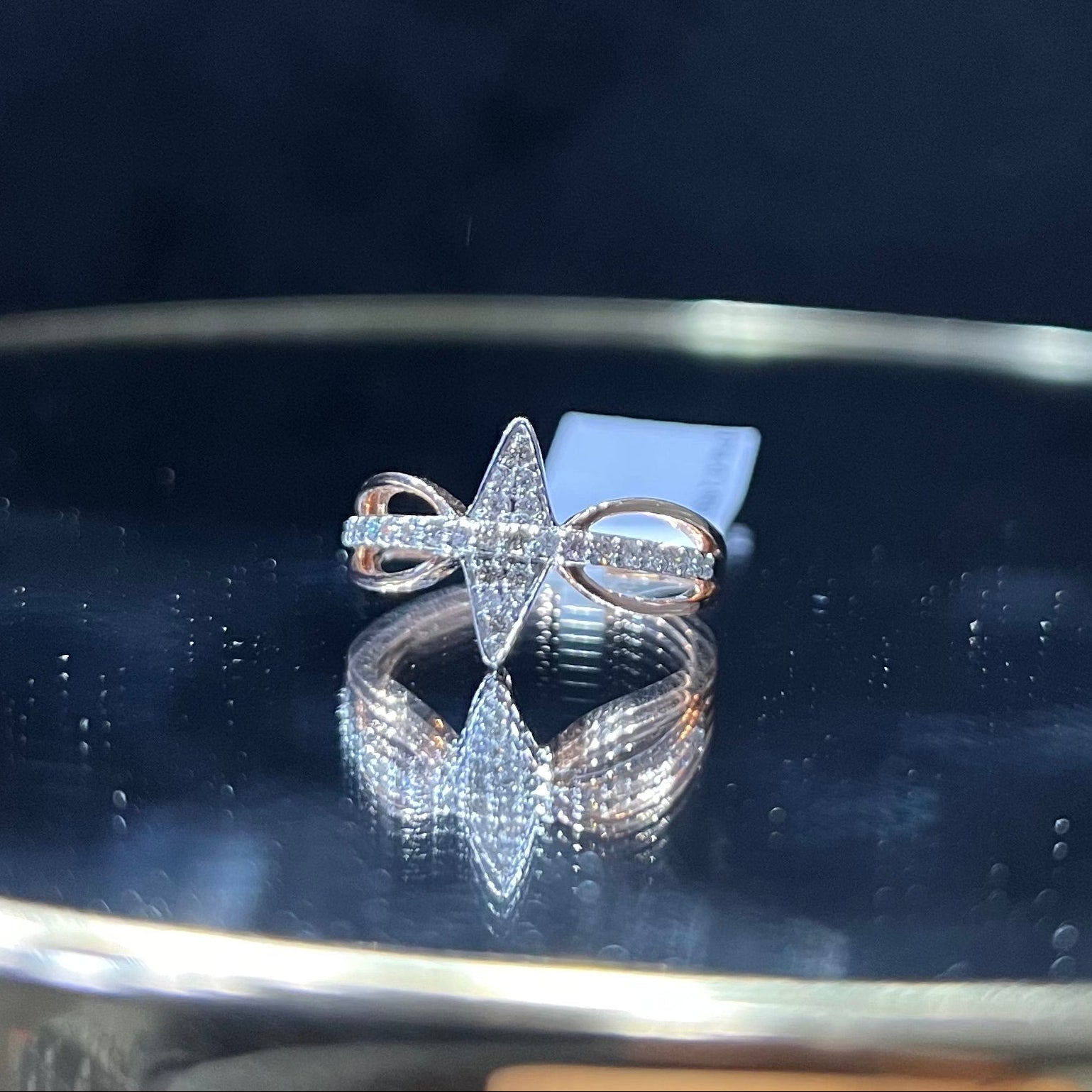 A handcrafted natural diamond solid gold ring, displayed on a reflective surface, featuring a brilliant cut diamond on a band with a star-shaped detail on the top.