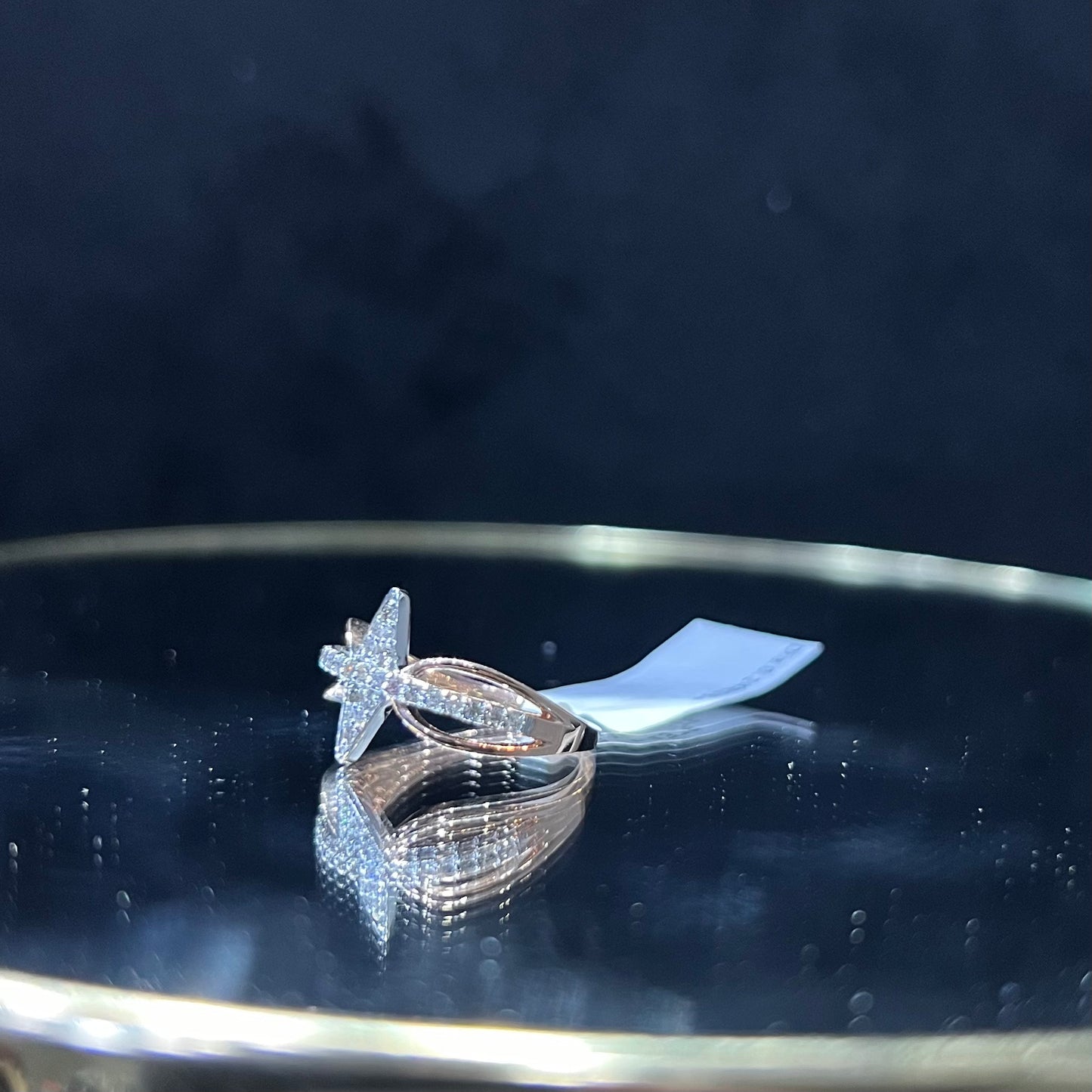 A handcrafted natural diamond solid gold ring, displayed on a reflective surface, featuring a brilliant cut diamond on a band with a star-shaped detail on the top.