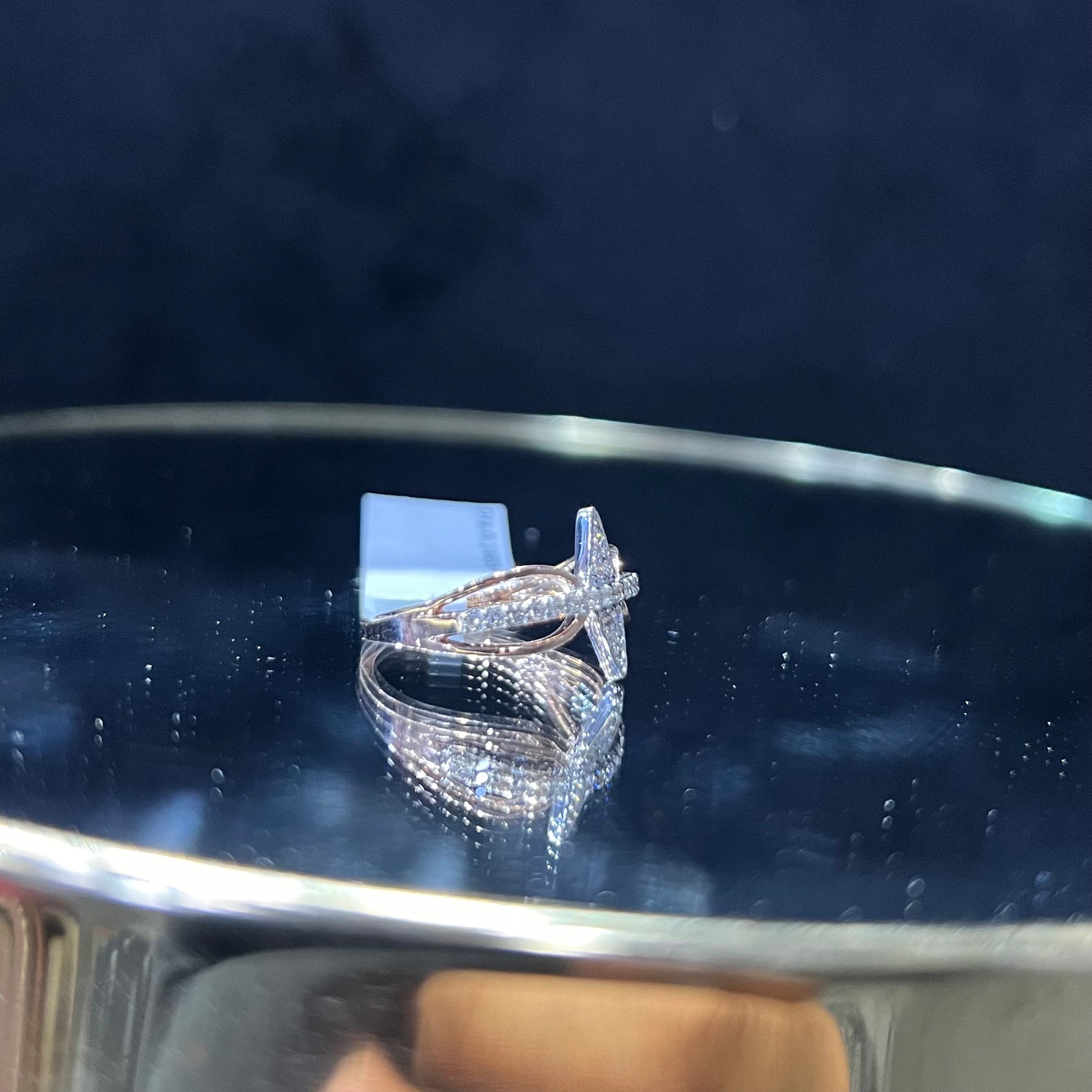 A handcrafted natural diamond solid gold ring, displayed on a reflective surface, featuring a brilliant cut diamond on a band with a star-shaped detail on the top.