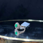 A handcrafted natural diamond solid gold ring, featuring a brilliant cut green stone centerpiece, surrounded by smaller clear diamonds, displayed on a reflective surface.