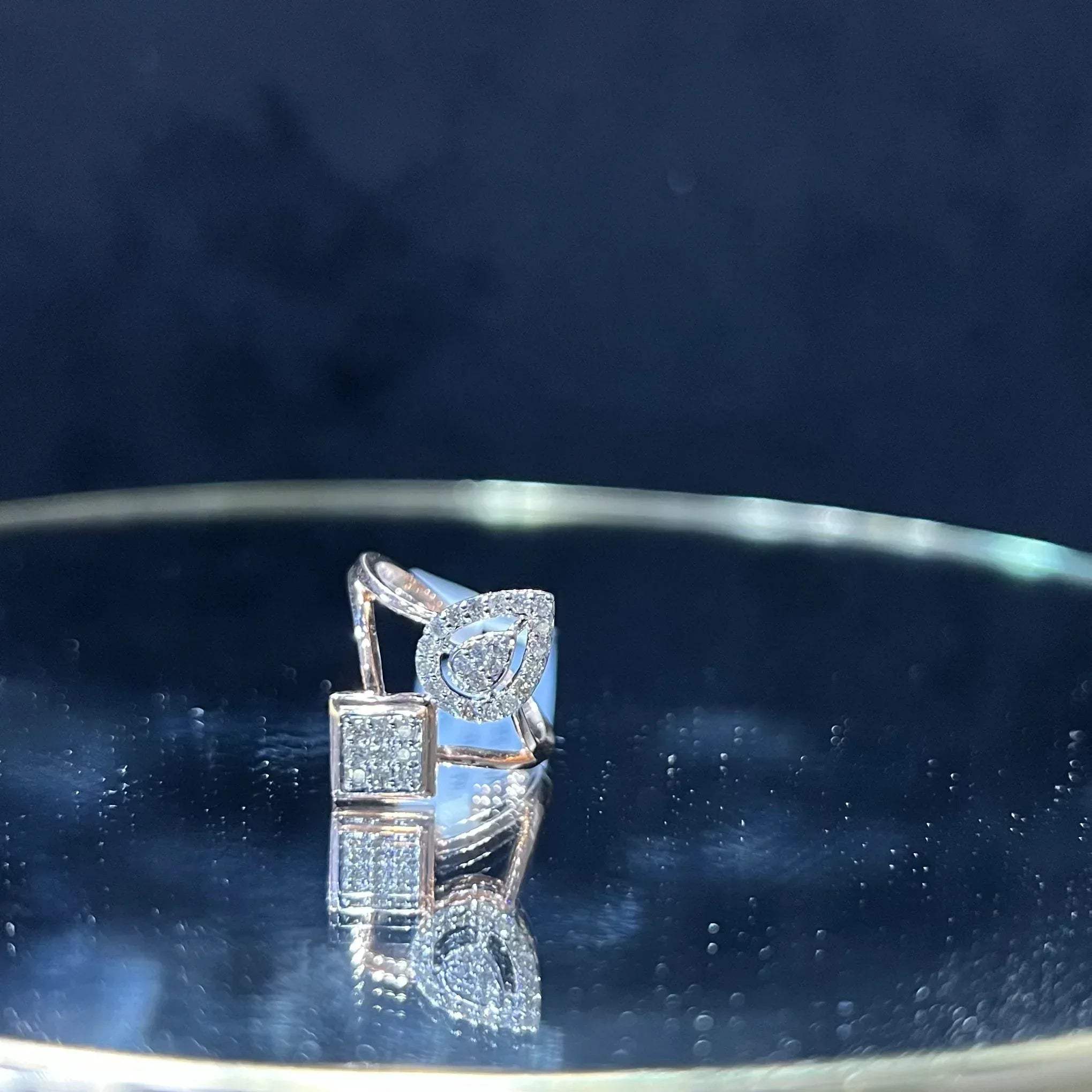 Elegant open-style diamond ring featuring a pear-shaped halo on one side and a square cluster on the other, displayed on a reflective metallic surface.