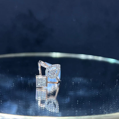 Elegant open-style diamond ring featuring a pear-shaped halo on one side and a square cluster on the other, displayed on a reflective metallic surface.