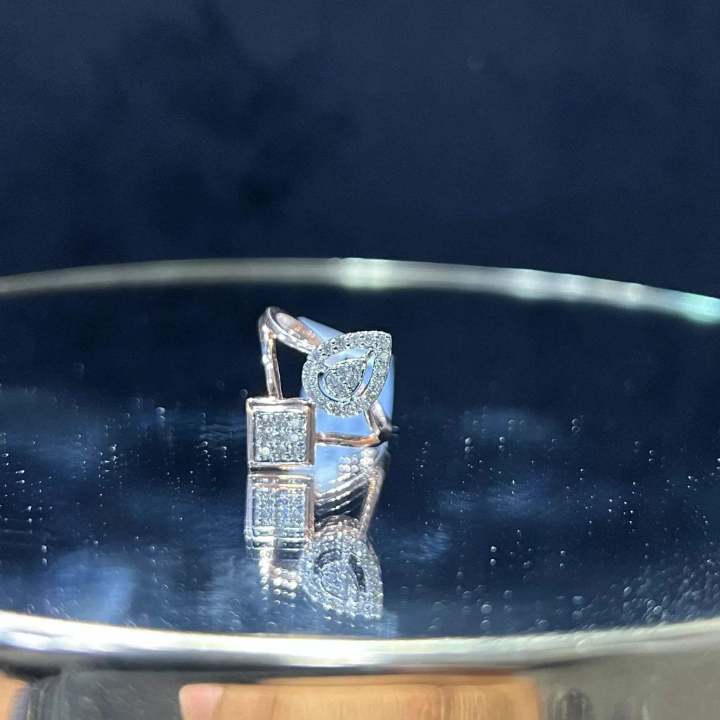 Elegant open-style diamond ring featuring a pear-shaped halo on one side and a square cluster on the other, displayed on a reflective metallic surface.