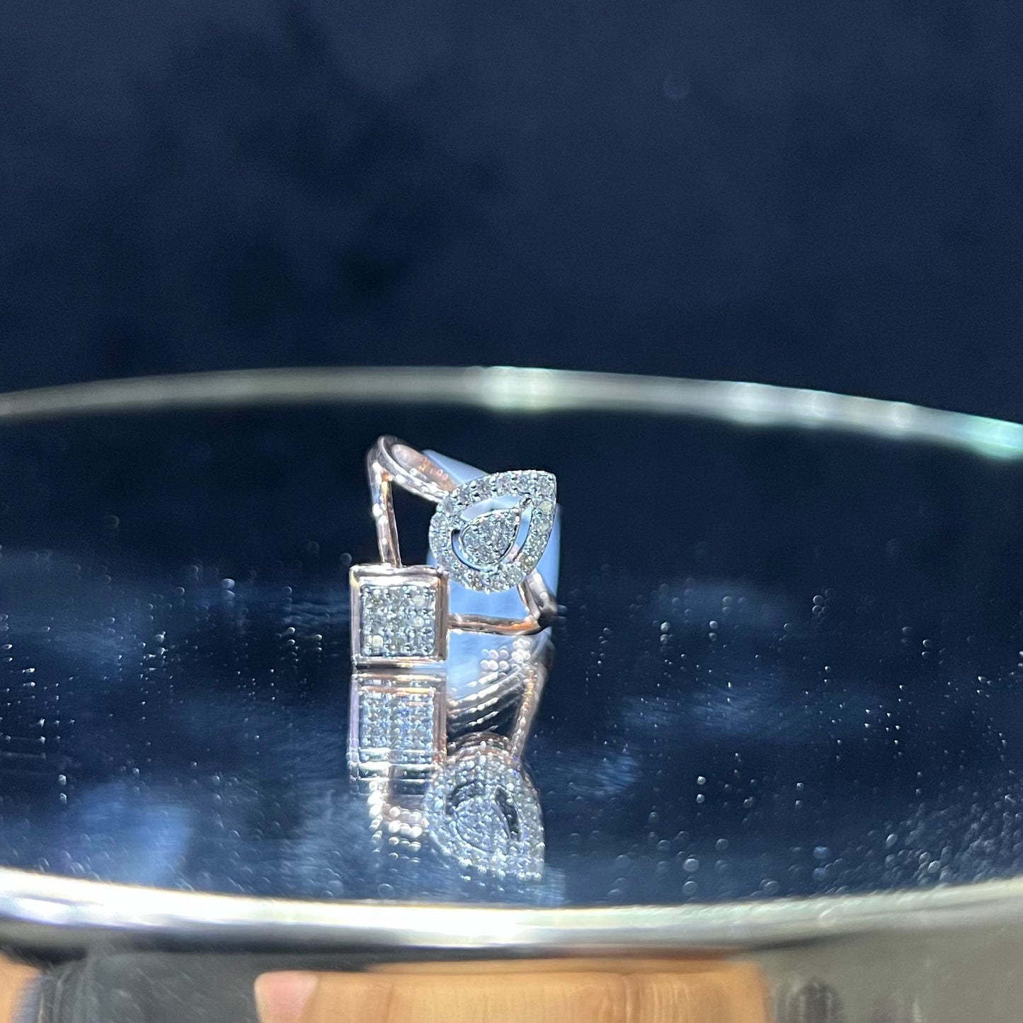 Elegant open-style diamond ring featuring a pear-shaped halo on one side and a square cluster on the other, displayed on a reflective metallic surface.