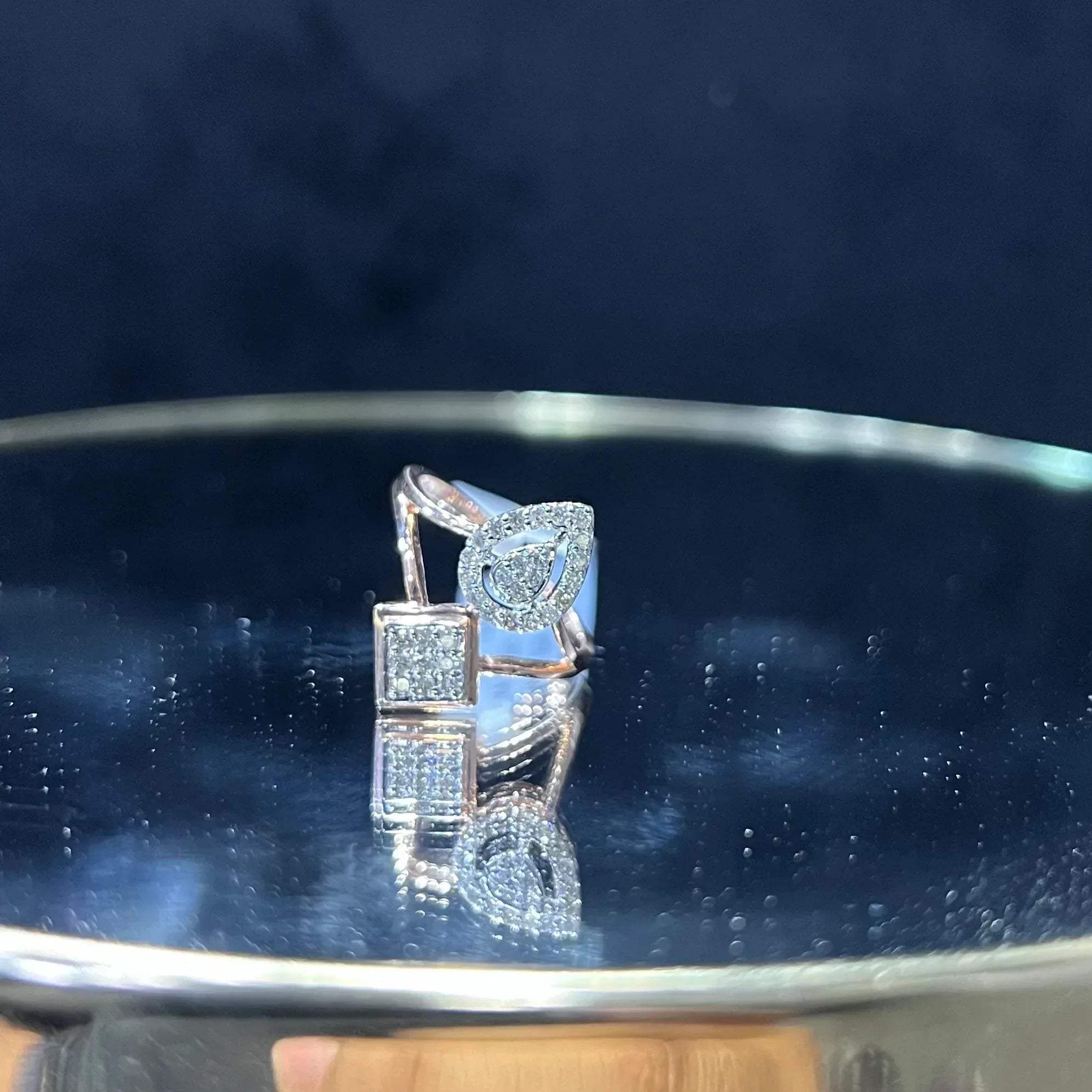 Elegant open-style diamond ring featuring a pear-shaped halo on one side and a square cluster on the other, displayed on a reflective metallic surface.