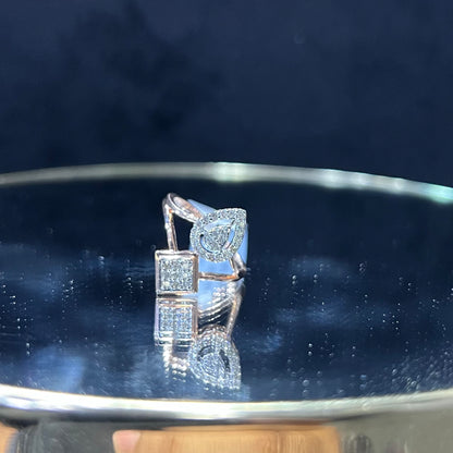 Elegant open-style diamond ring featuring a pear-shaped halo on one side and a square cluster on the other, displayed on a reflective metallic surface.