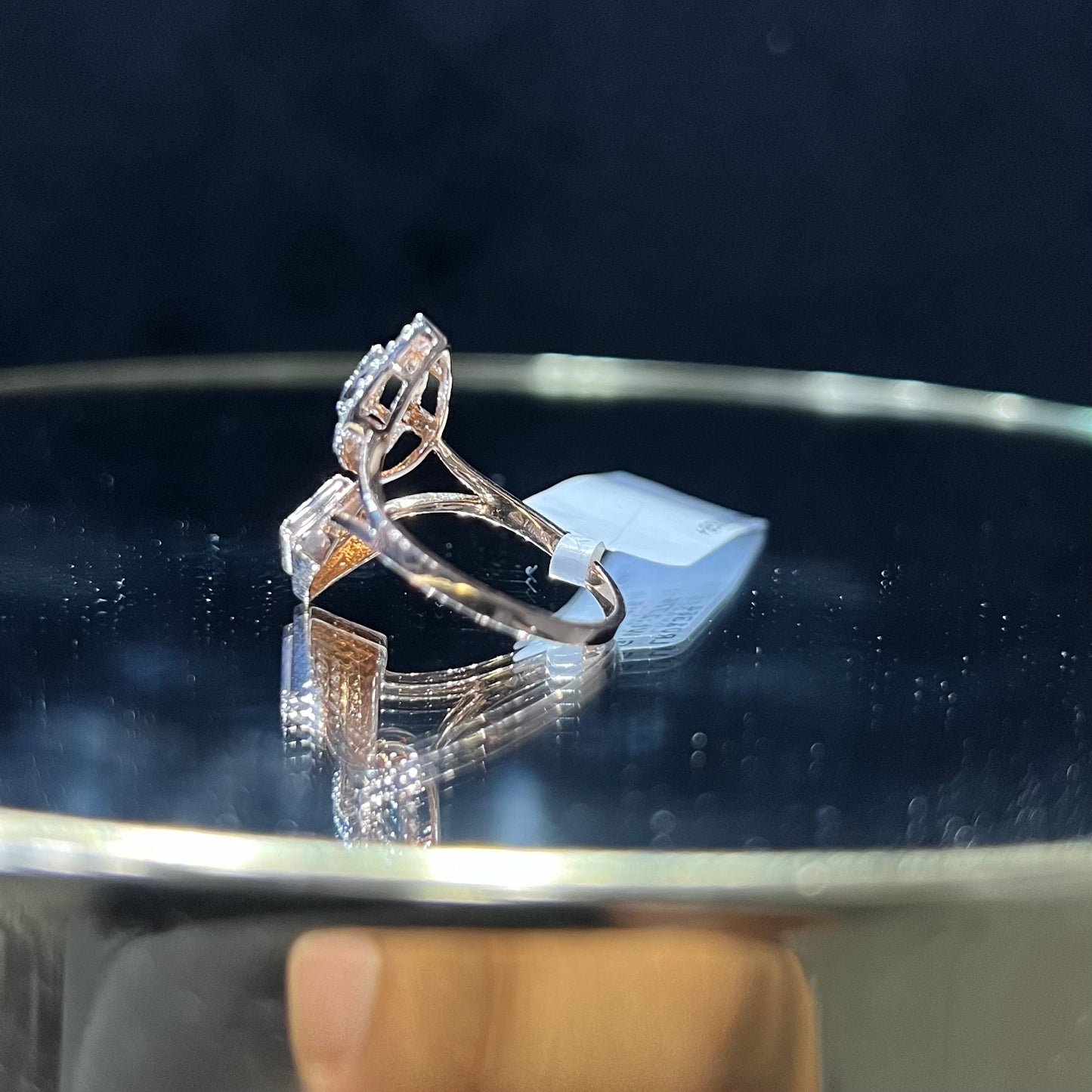 Elegant open-style diamond ring featuring a pear-shaped halo on one side and a square cluster on the other, worn in a black mannequin's hand displayed displayed against a reflective metallic background.