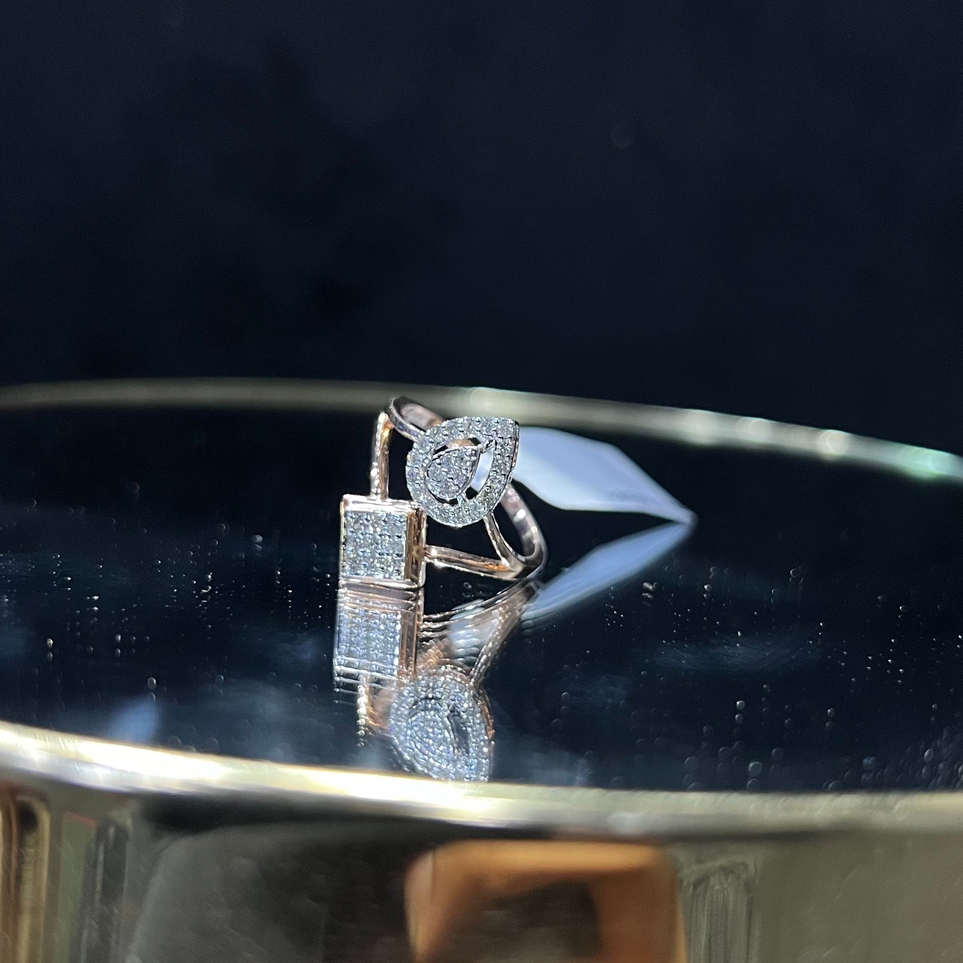 Elegant open-style diamond ring featuring a pear-shaped halo on one side and a square cluster on the other, worn in a black mannequin's hand displayed displayed against a reflective metallic background.