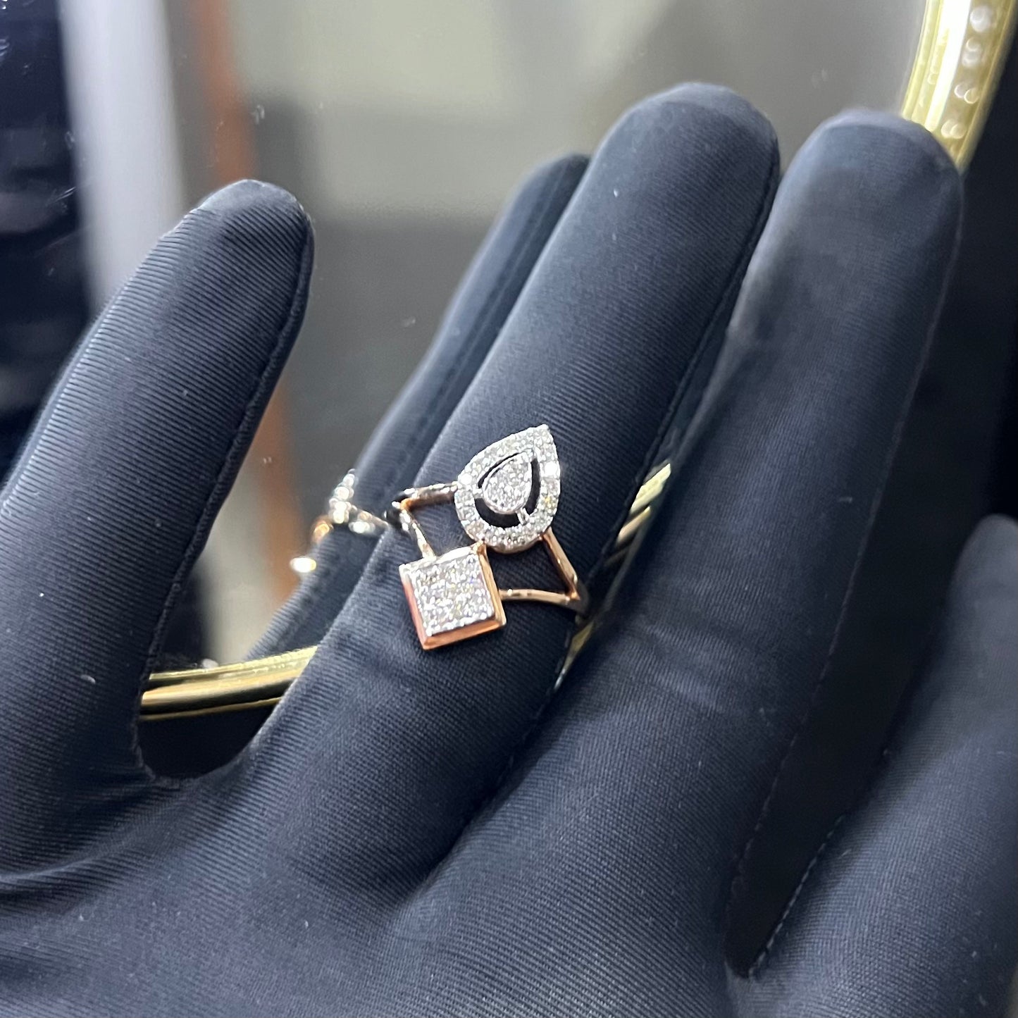 Elegant open-style diamond ring featuring a pear-shaped halo on one side and a square cluster on the other, worn in a black mannequin's hand displayed displayed against a reflective metallic background.