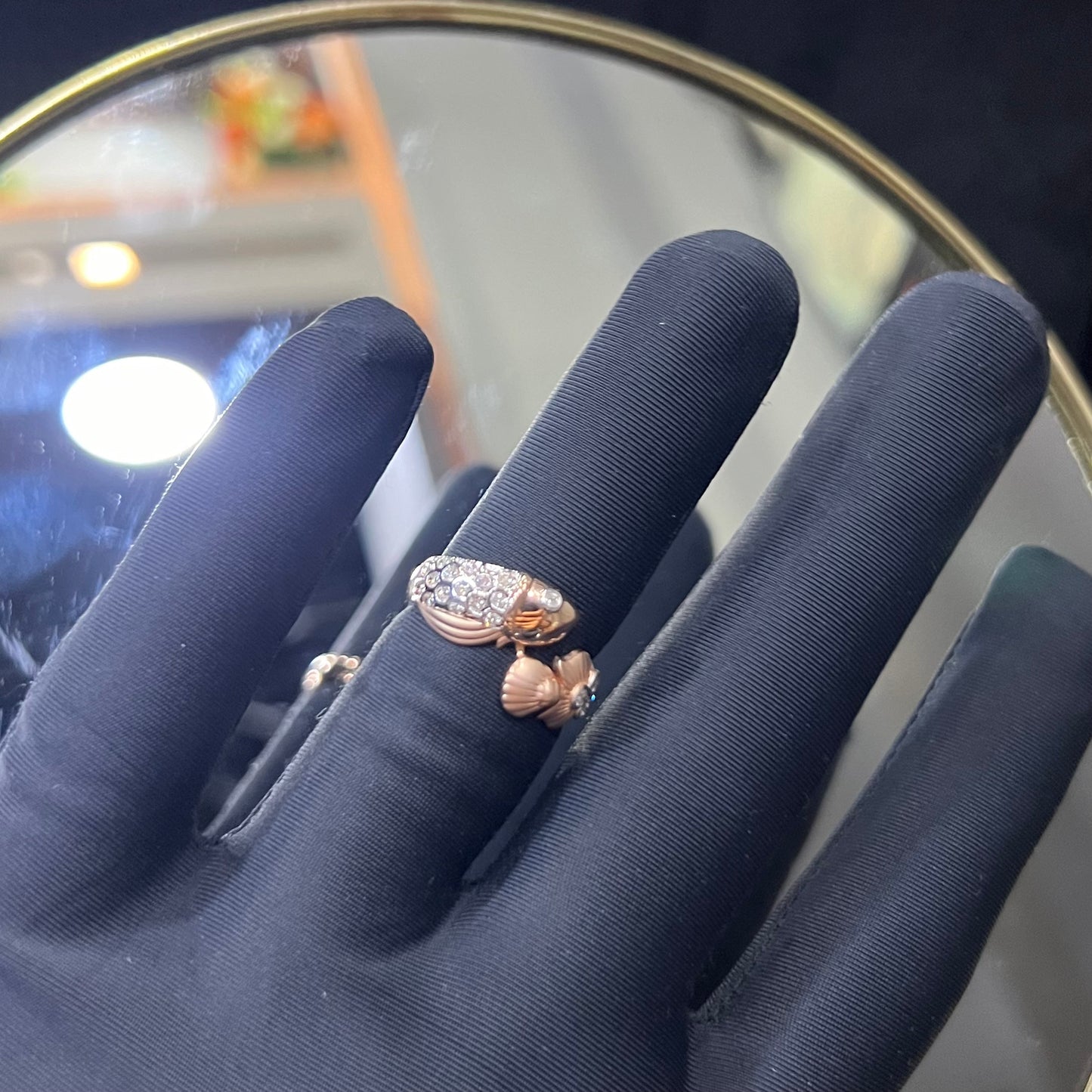 A ring with a gold band and a central natural diamonds, held by a hand with a black glove, with a reflective surface in the background.