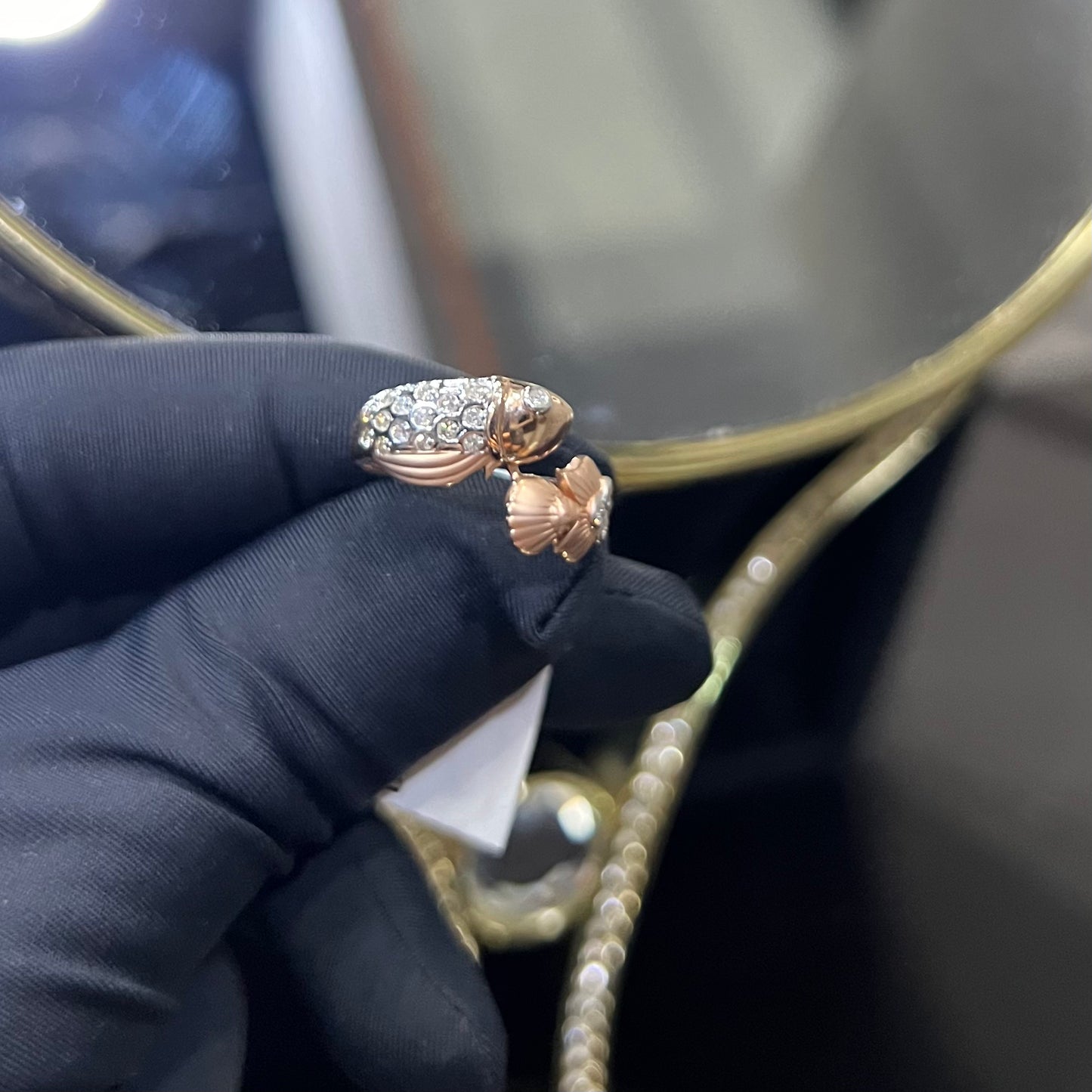 A ring with a gold band and a central natural diamonds, held by a hand with a black glove, with a reflective surface in the background.