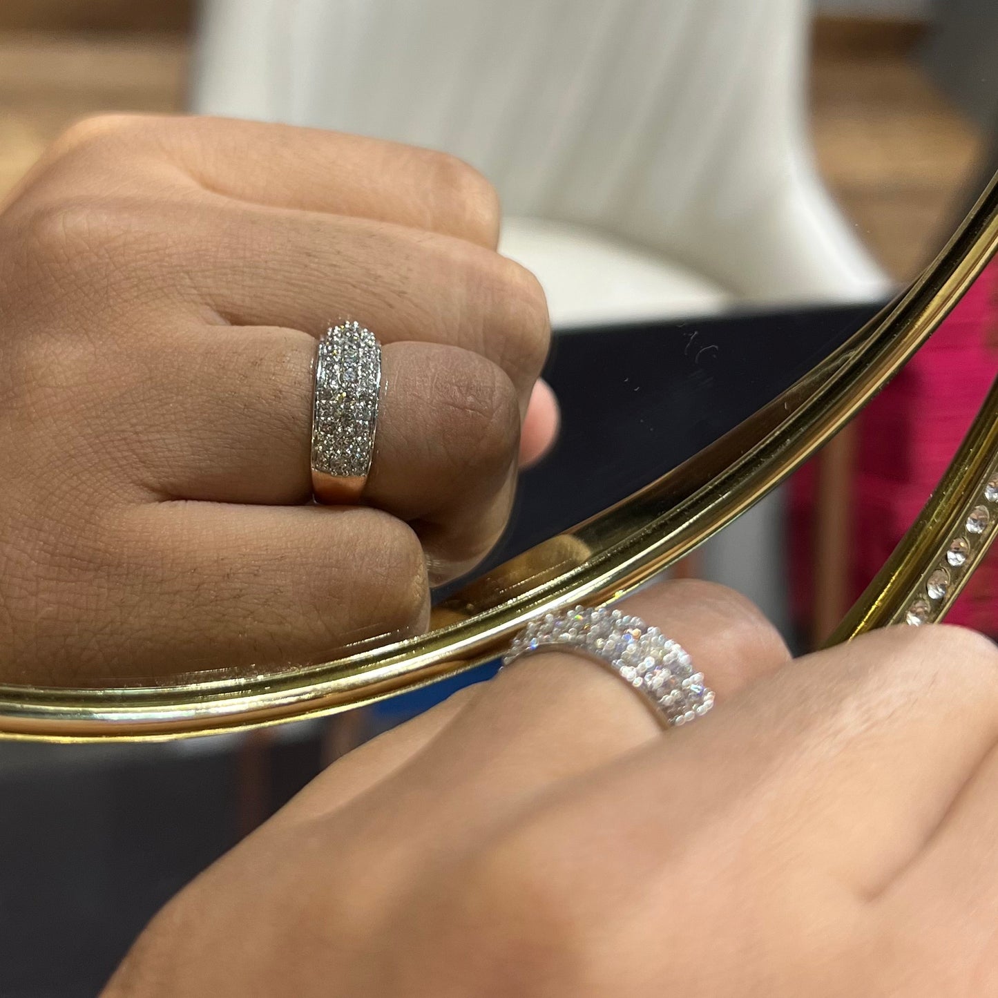 Close-up of a hand wearing a wide diamond band ring, featuring multiple rows of sparkling round-cut diamonds set in gold, reflected elegantly in a gold-framed mirror.