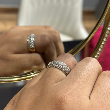 Close-up of a hand wearing a wide diamond band ring, featuring multiple rows of sparkling round-cut diamonds set in gold, reflected elegantly in a gold-framed mirror.