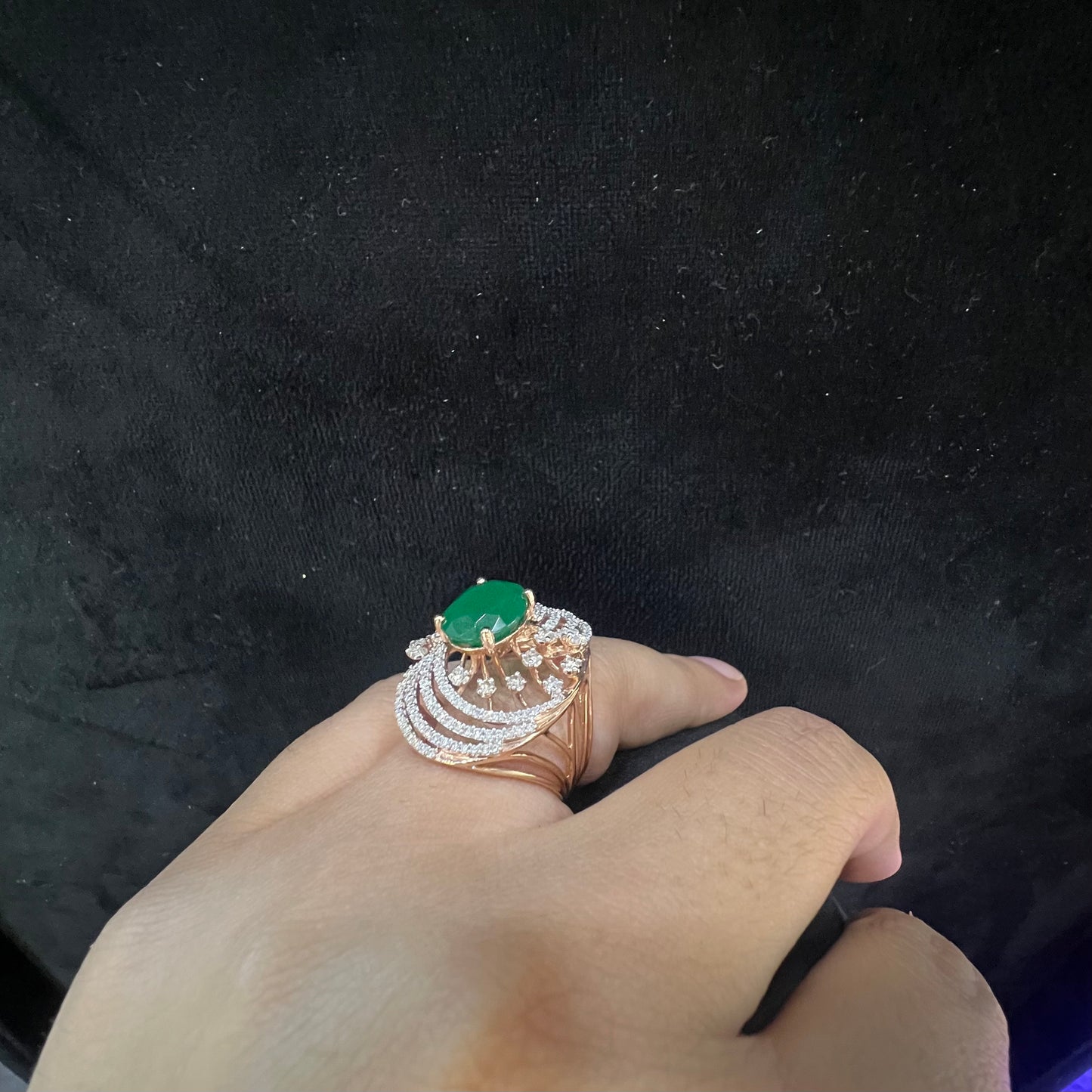 Close-up of a hand holding an elegant statement ring featuring a large oval green gemstone at the center, surrounded by concentric circles of sparkling white diamonds set in a gold base against a black background.