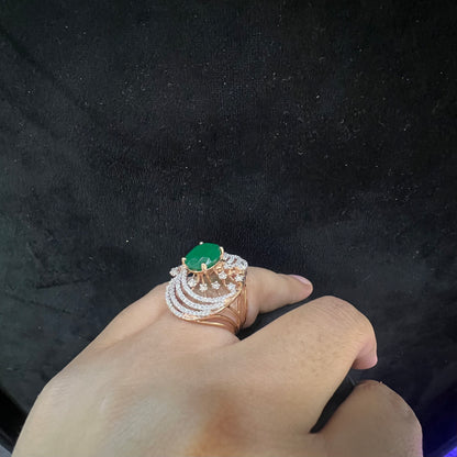 Close-up of a hand holding an elegant statement ring featuring a large oval green gemstone at the center, surrounded by concentric circles of sparkling white diamonds set in a gold base against a black background.