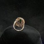 Close-up of a an elegant statement ring featuring a large oval green gemstone at the center, surrounded by concentric circles of sparkling white diamonds set in a gold base against a black background.