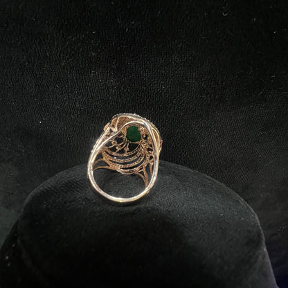 Close-up of a an elegant statement ring featuring a large oval green gemstone at the center, surrounded by concentric circles of sparkling white diamonds set in a gold base against a black background.